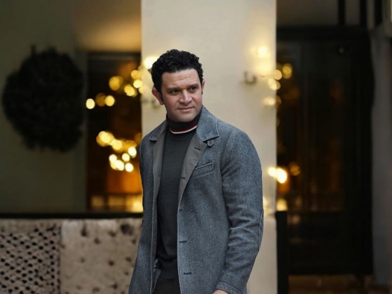 A man smiles outdoors in stylish clothing.