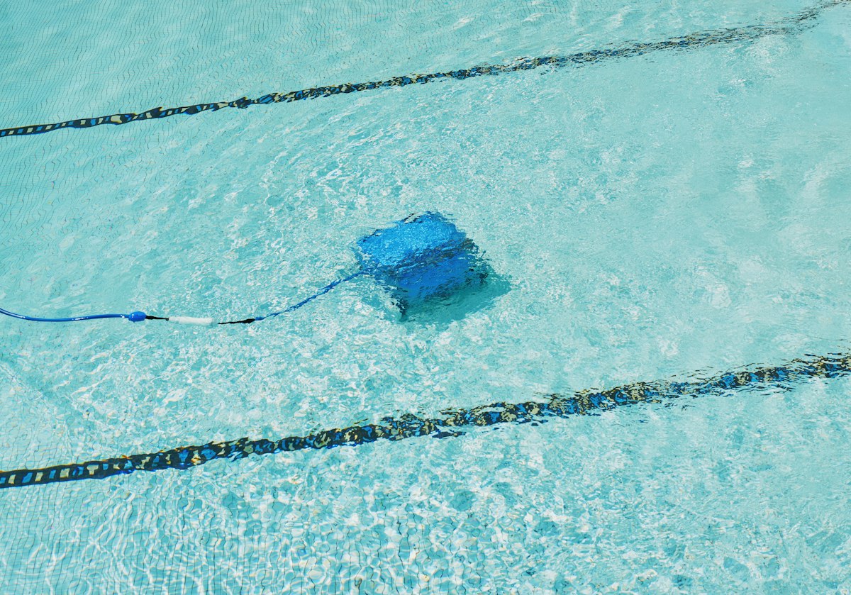 Robot limpiafondos azul con su cable trabajando en el fondo de una pileta de agua celeste y cristalina con líneas de carril