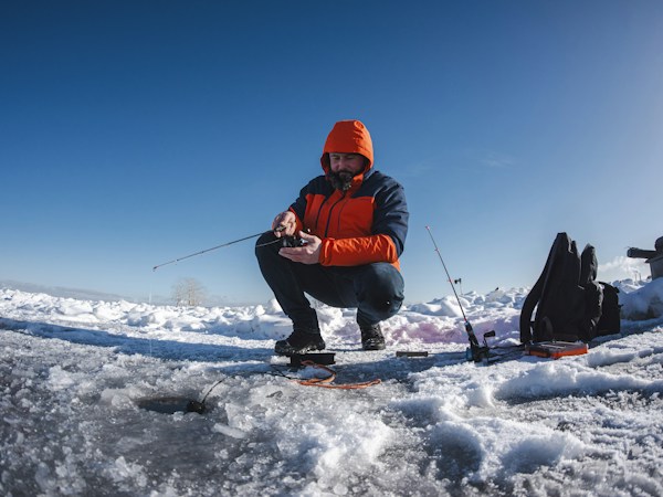 Ice angler in an orange hooded jacket crouching bare-handed over a hole in a frozen lake, working a short rod under a clear blue sky
