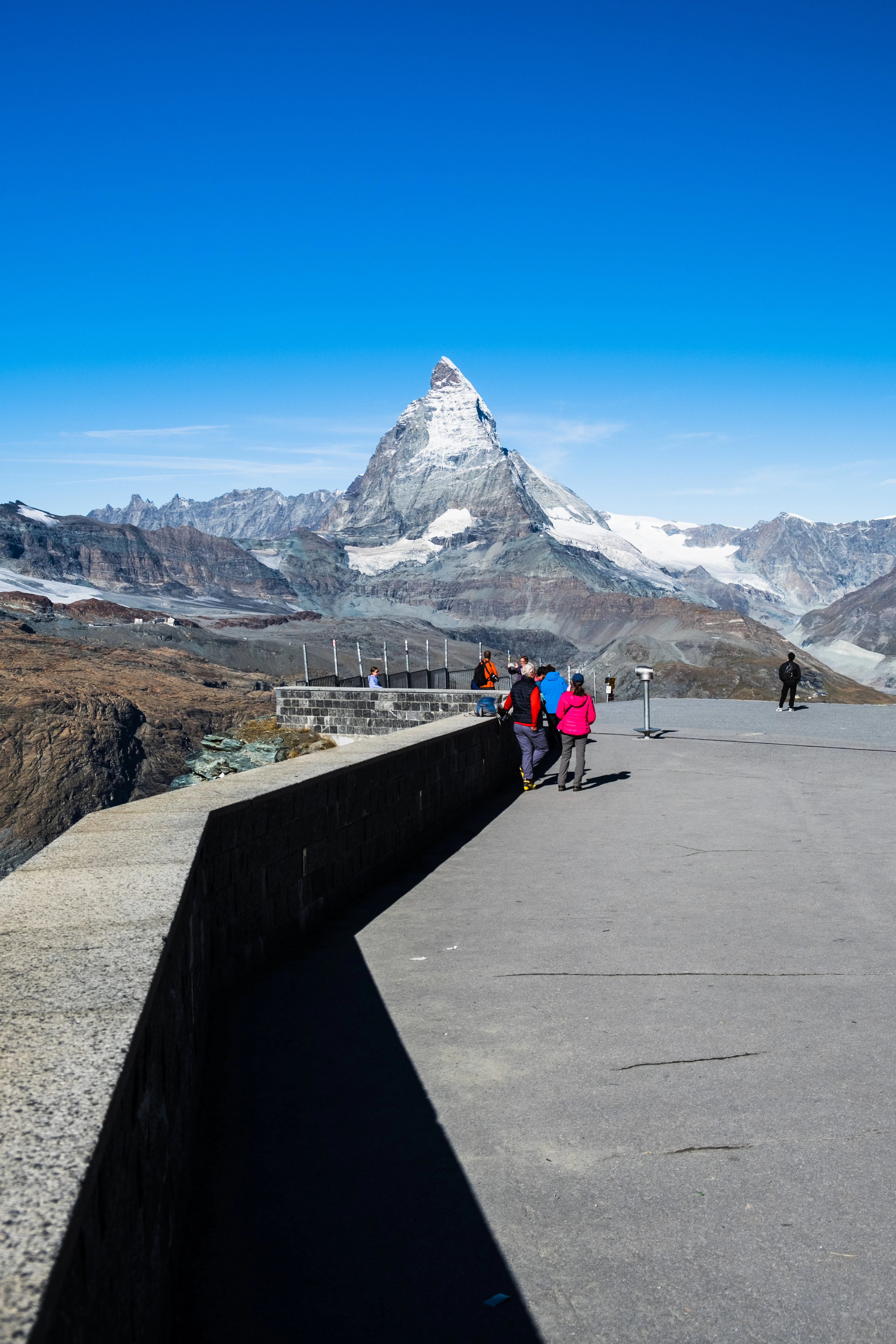 Visitors stand on a stone viewing terrace admiring the Matterhorn under a clear blue sky.