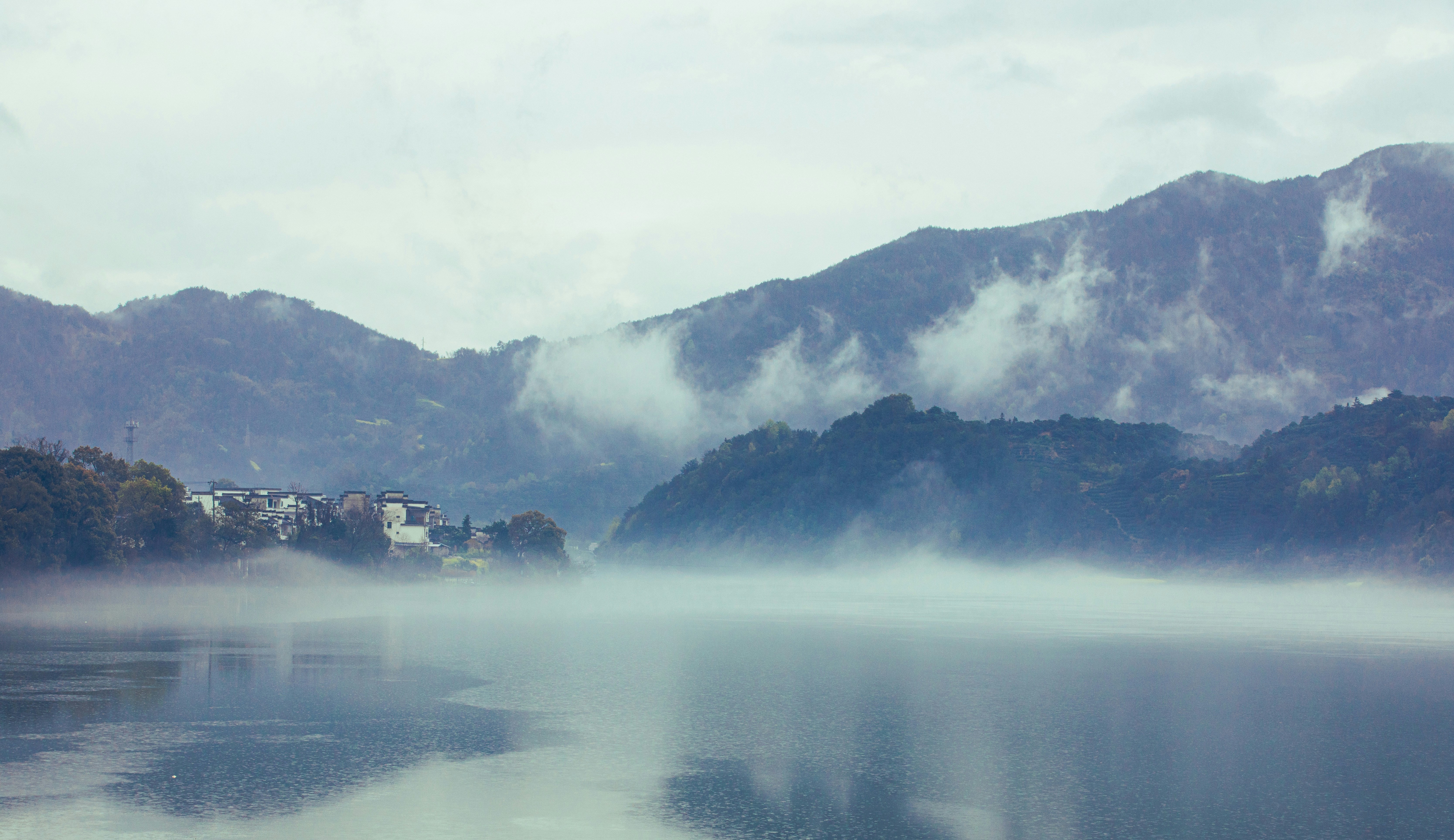 Foggy lake reflects mountain range on a cloudy day.