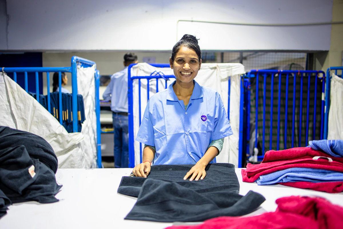 Une employée en chemise de travail bleu clair plie une serviette sombre sur une table de tri, devant des chariots métalliques bleus et des piles de linge de couleur dans un atelier de blanchisserie