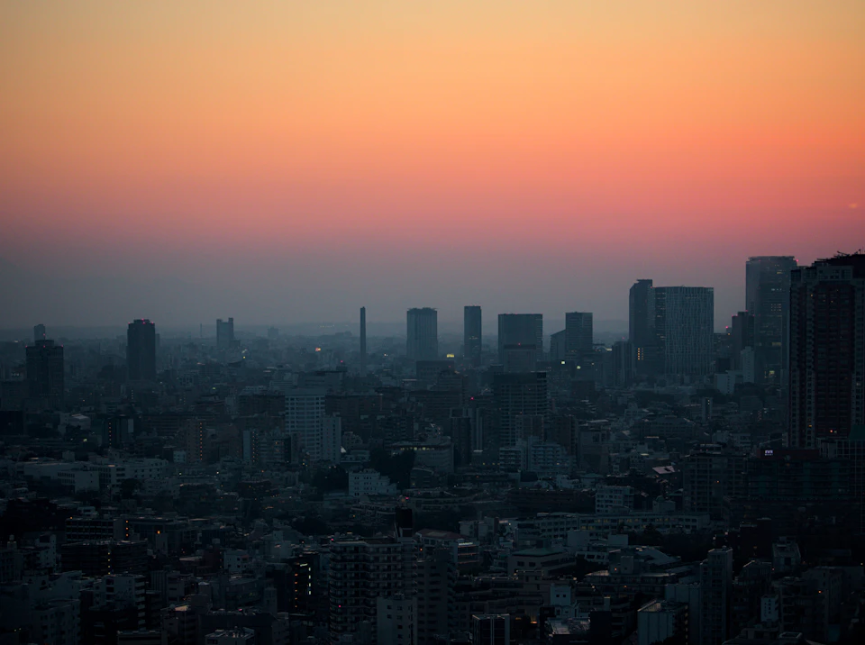 The image presents a panoramic view of a cityscape at sunset, with the sky painted in hues of orange and pink. The sun is setting behind the buildings, casting long shadows across the urban landscape. The city's skyline features tall buildings of varying heights, their windows reflecting the warm glow of the setting sun. The perspective from which the photo was taken allows for a comprehensive view of the city below, showcasing its architectural diversity and the interplay between nature and human-made structures.

The sky is filled with clouds, adding depth to the scene and creating a sense of vastness in the image. The colors of the sunset are vibrant and striking, enhancing the overall visual appeal of the photograph.