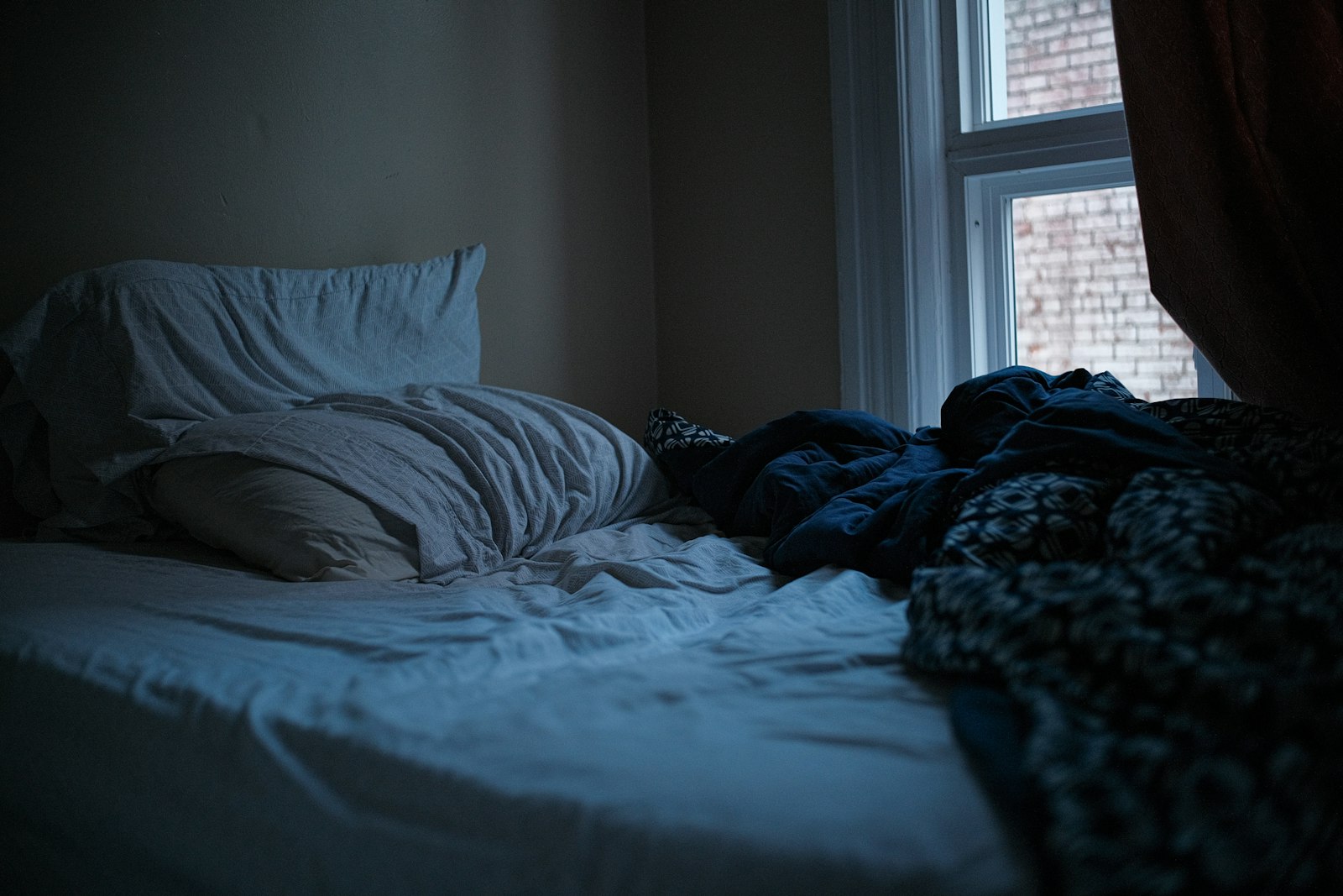 Unmade bed in a dark quiet bedroom, dawn light — the recovery a 24/7 wearable measures
