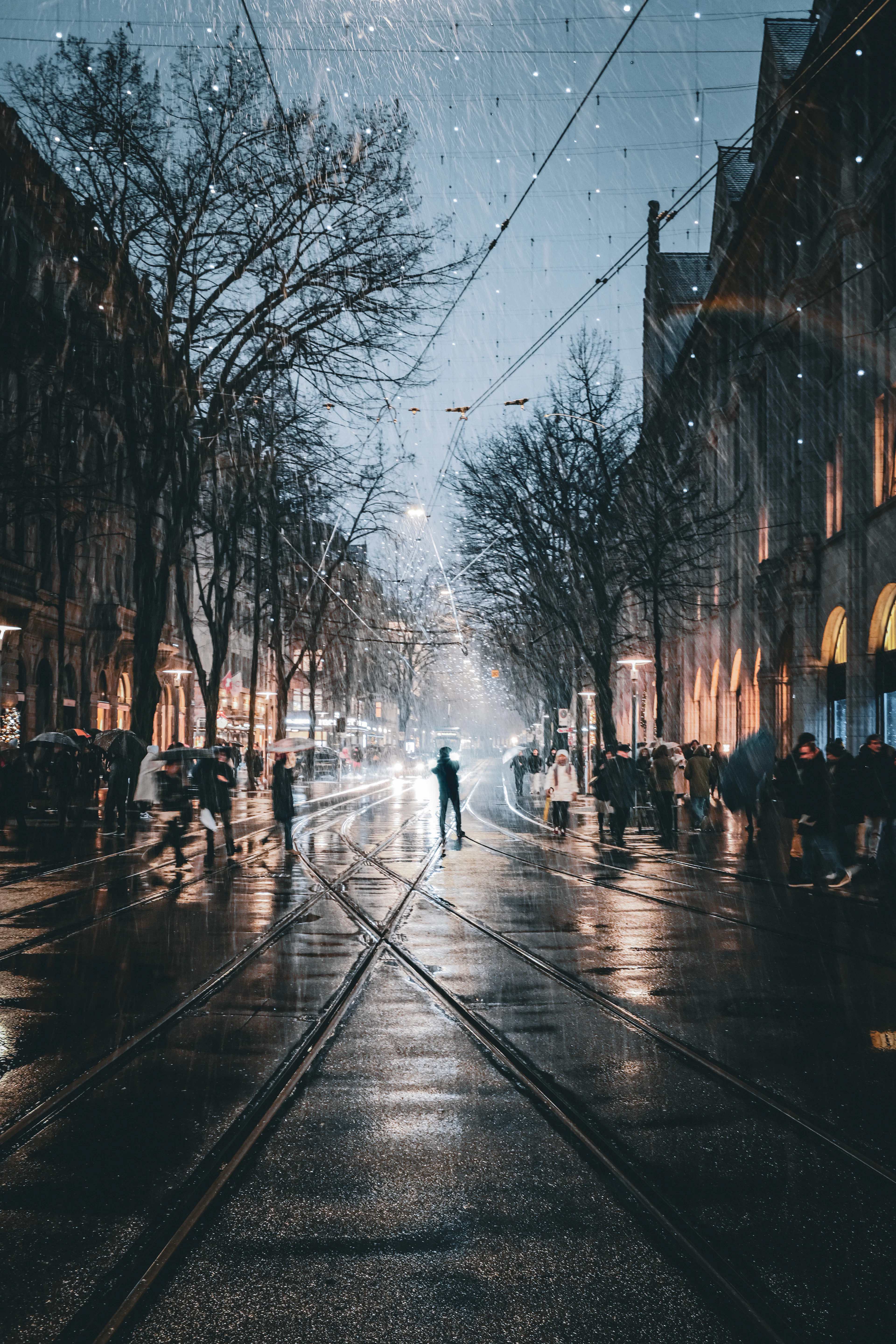 Rainy night street with glistening tram tracks, string lights overhead, and pedestrians carrying umbrellas.