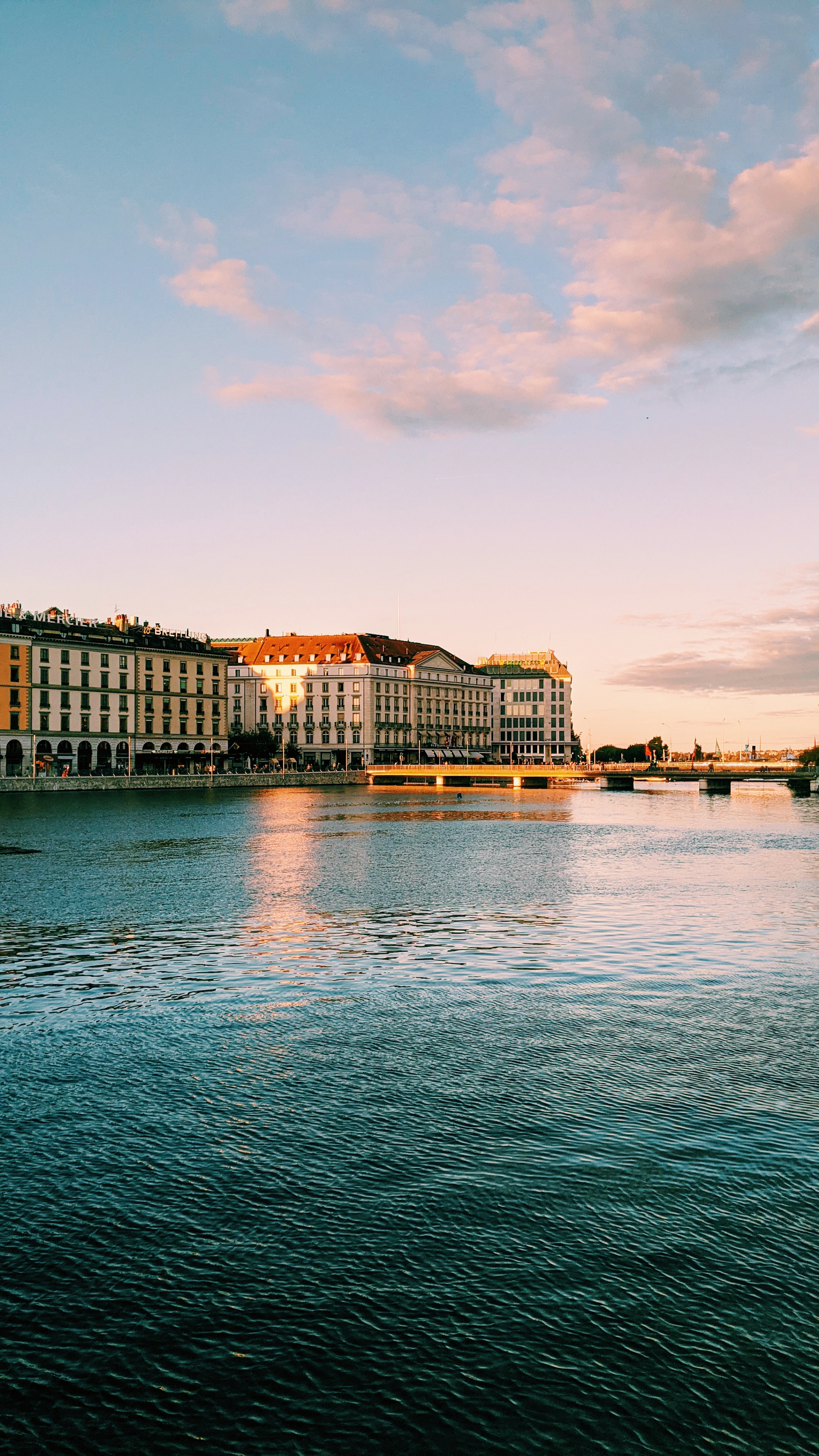 Geneva's waterfront at golden hour seen from the water, the long line of pale nineteenth-century hotel and cafe facades along the quay catching the last sun beneath a pink-streaked sky