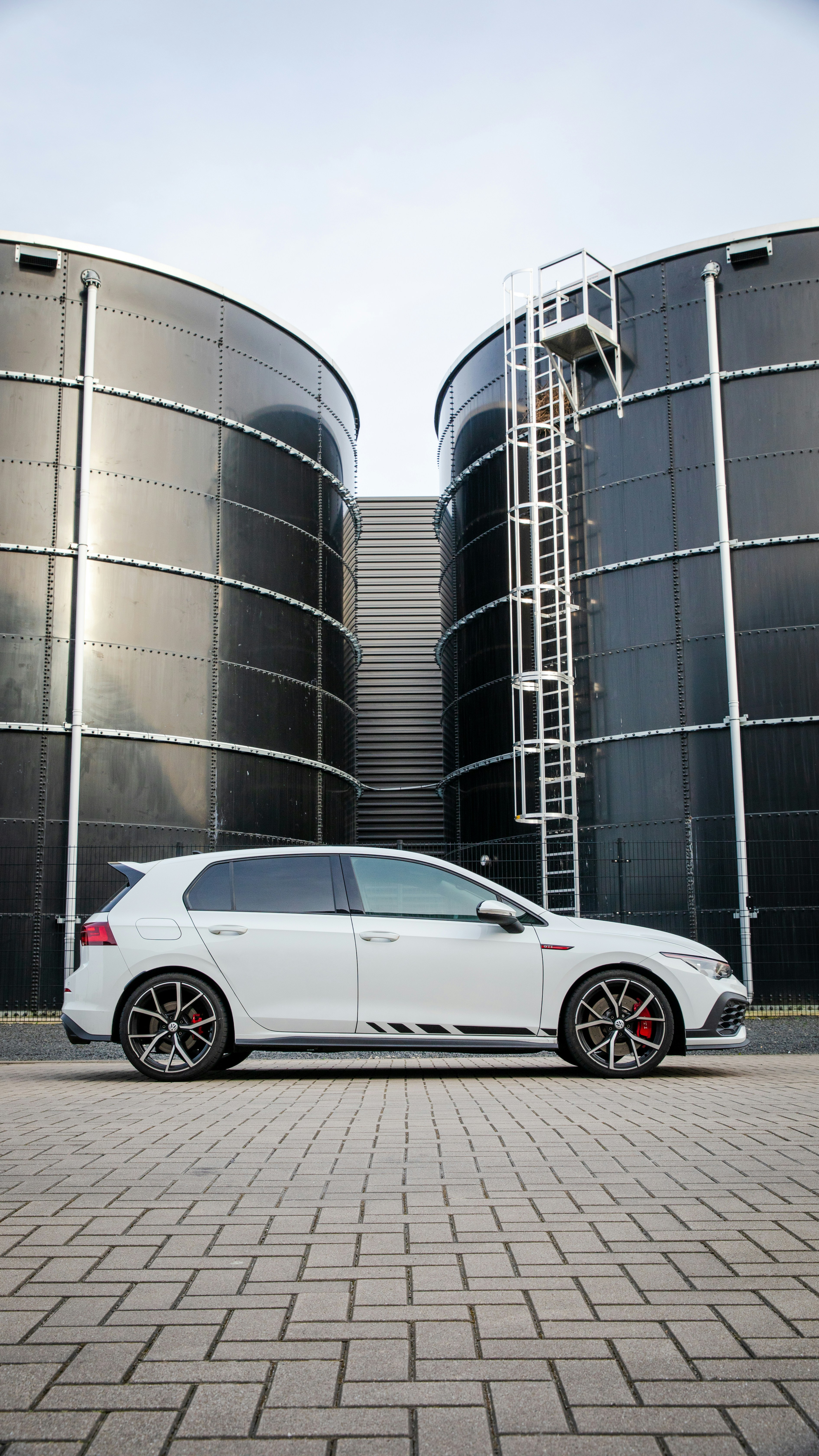 A white car is parked beside industrial tanks.