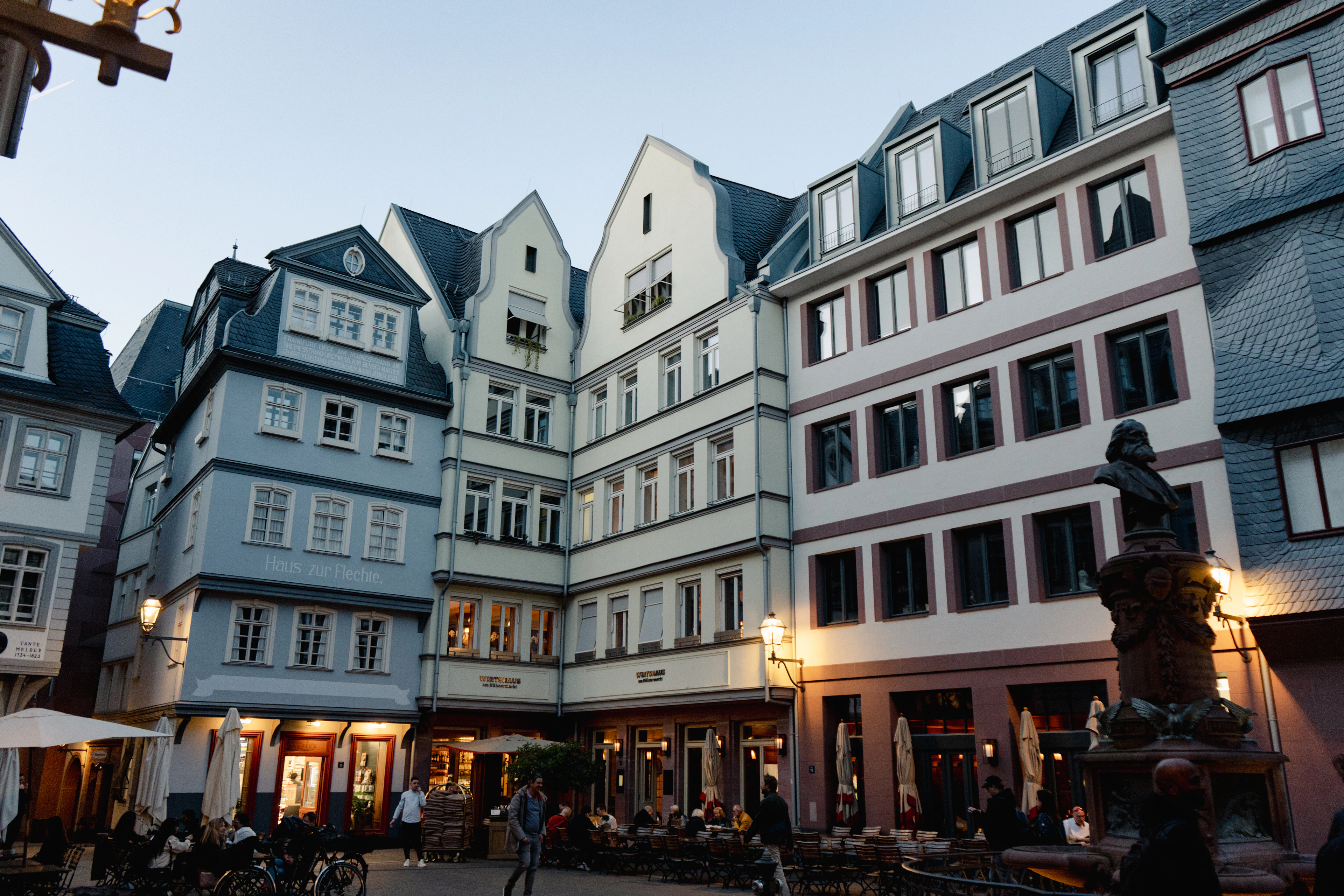 Restored gabled townhouses in pale blue, cream and pink around a square in Frankfurt's Neue Altstadt at blue hour, their ground-floor cafe windows lit and a bronze statue on a stone plinth at the right