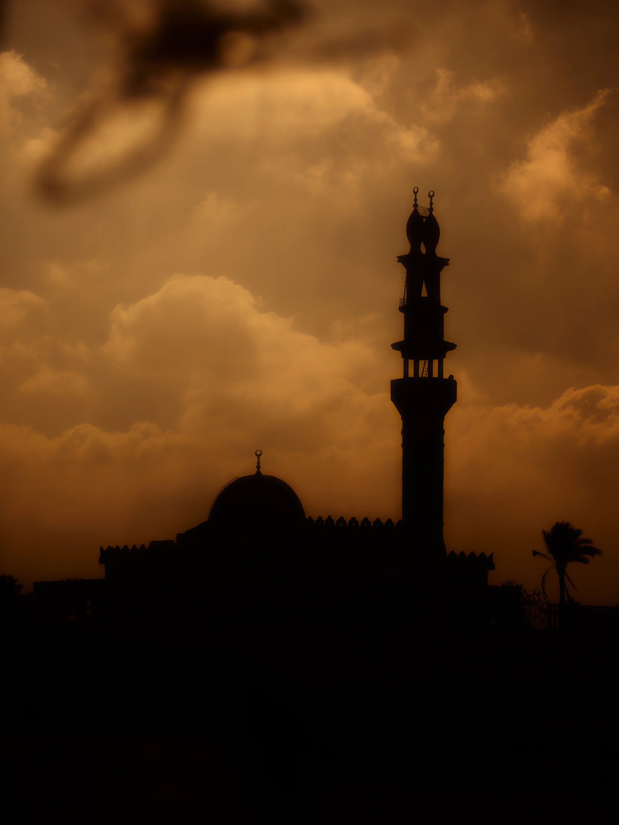 Silhouette of a mosque dome and a minaret topped with a crescent against an orange, cloudy sky