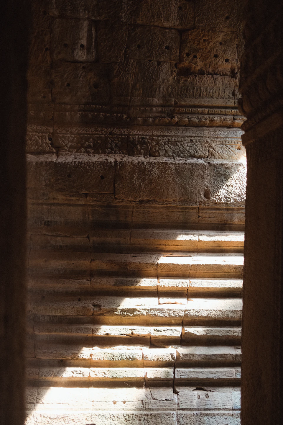 The image shows a stone wall with a series of steps leading up to it, bathed in sunlight streaming through an opening on the right side. The light casts long shadows and highlights the texture of the stone, creating a sense of depth and dimension. The perspective is from below, looking upwards towards the top of the wall, giving the impression that one is ascending the stairs.