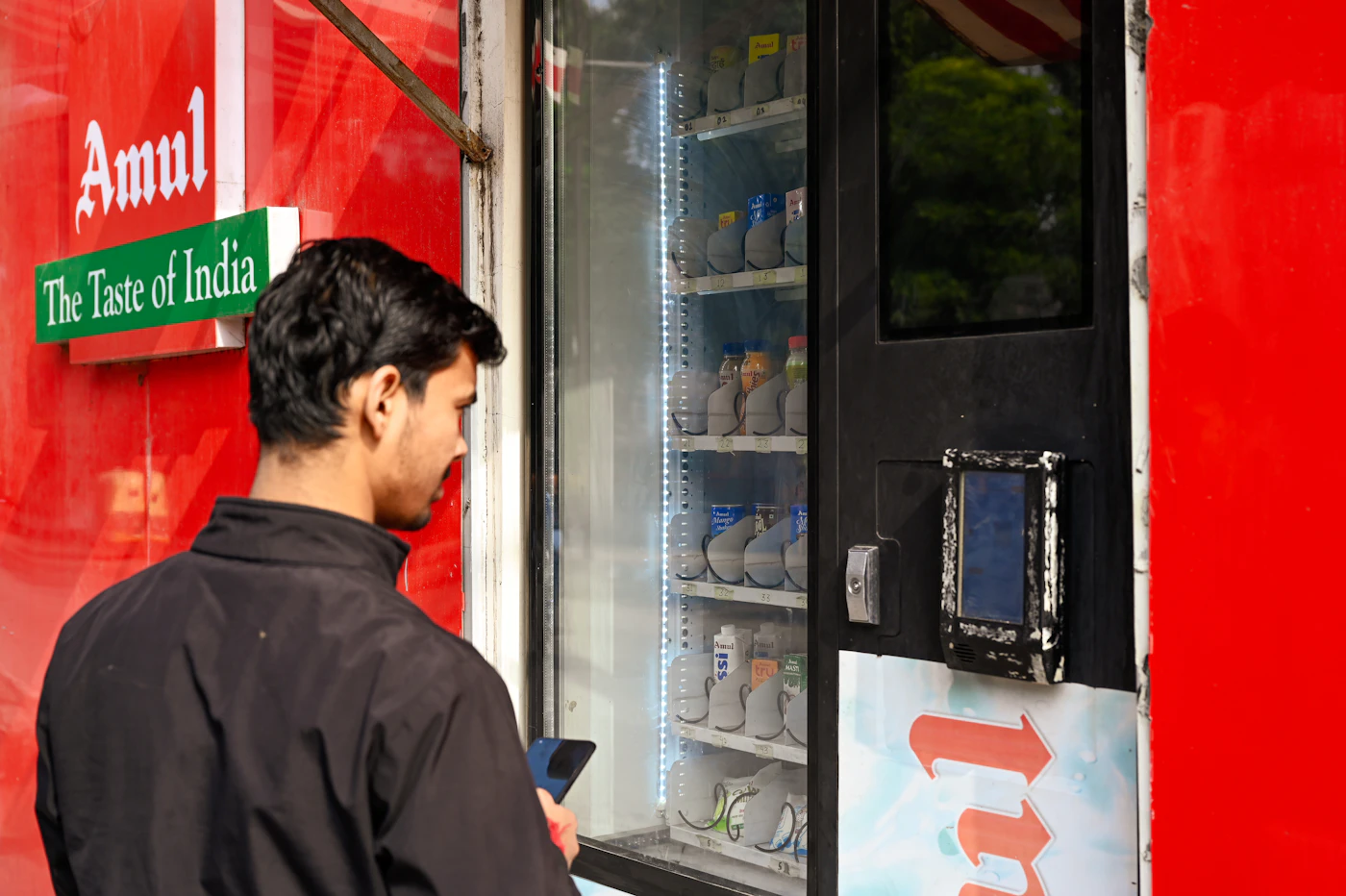 A young man holding a smartphone in front of an Amul vending machine
