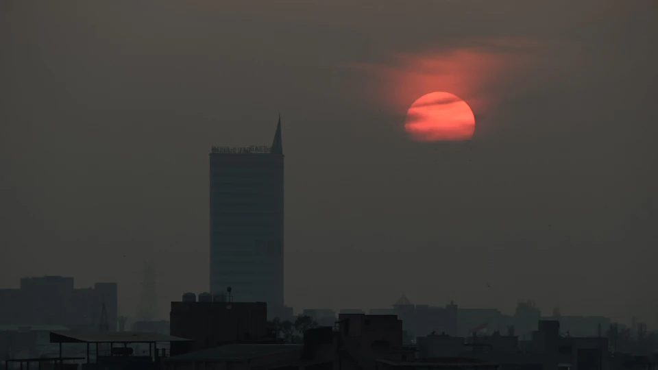 The image depicts a city skyline at sunset, with the sun setting behind the buildings and casting a warm glow over the scene. The sky is filled with orange hues from the setting sun, creating a stunning backdrop for the urban landscape below. In the foreground, there are several tall buildings of varying heights, including a large skyscraper that stands out against the skyline. The cityscape is bathed in the soft light of the sunset, and the image conveys a sense of tranquility and beauty as day transitions into night.

The perspective of the photo is from below, looking up at the towering buildings, which adds to the grandeur of the scene. The sun's position in the sky casts long shadows across the cityscape, adding depth and dimension to the image.