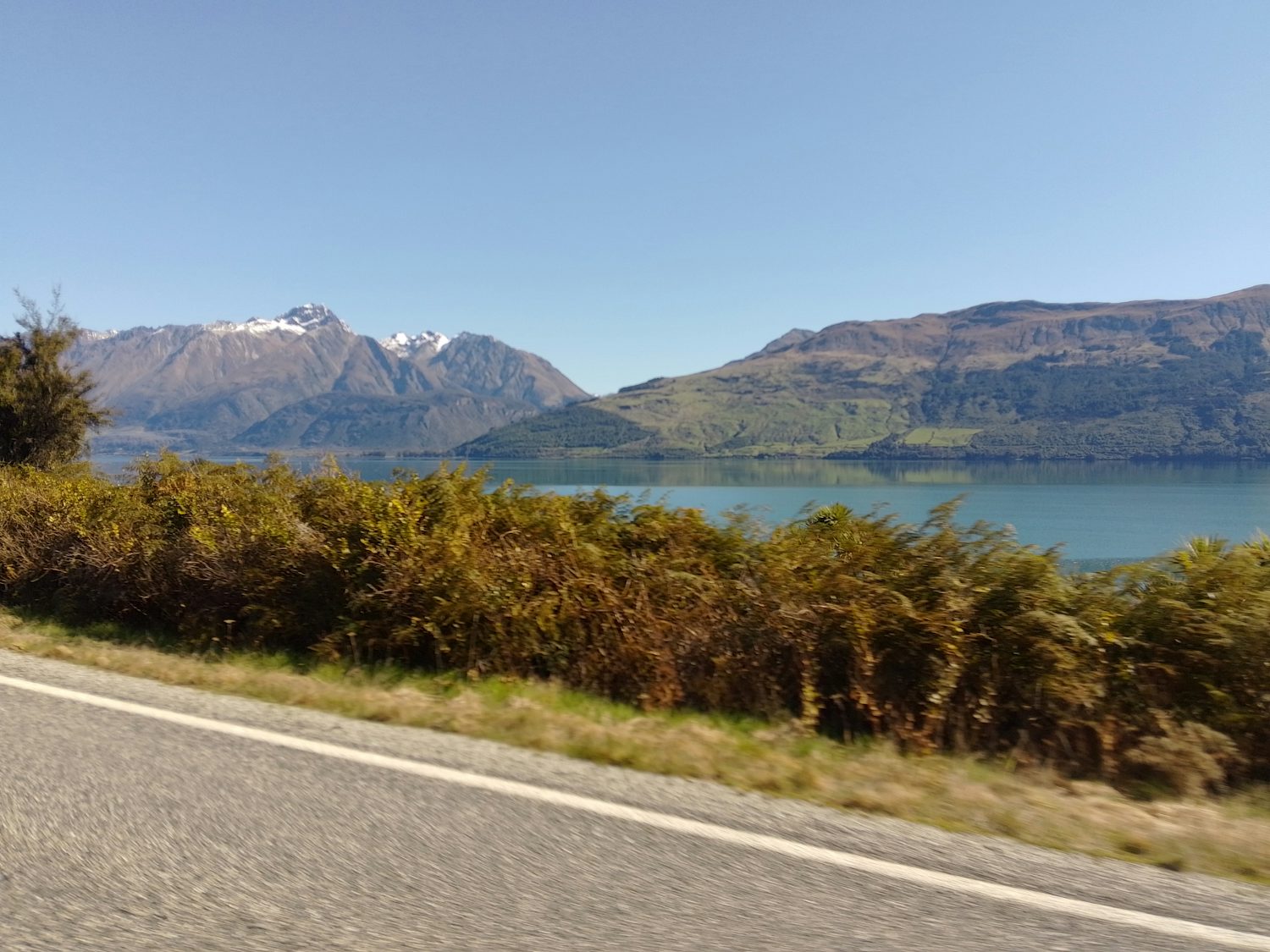 In the foreground is a road, with grass and shrubs edging it. Behind it is water that reflects mountains and the blue sky behind it. One of the mountains is taller and snow-capped; the other is more green.