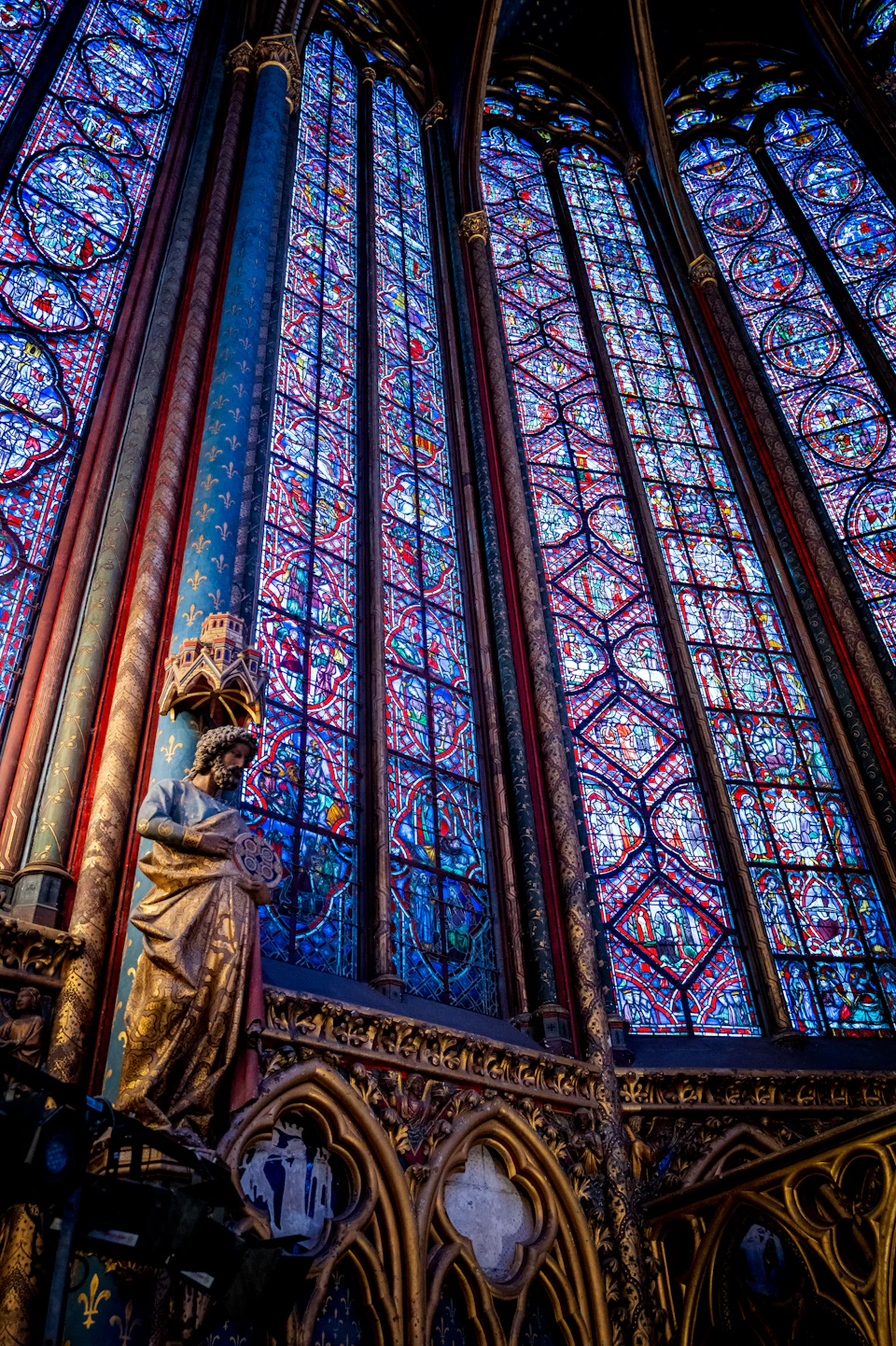 The image depicts a majestic cathedral with tall, pointed arches and stained glass windows that fill the interior with vibrant colors. The perspective is from below, looking up at the towering structure. A statue stands prominently on the left side of the cathedral, adding to its grandeur. The cathedral's architecture features intricate details and patterns, showcasing the craftsmanship involved in its construction.