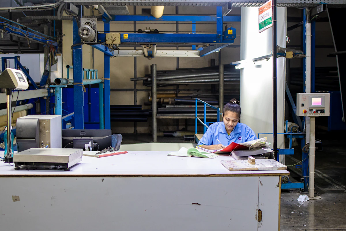 A woman in a blue work uniform goes through ledgers at a desk on a factory floor surrounded by machinery