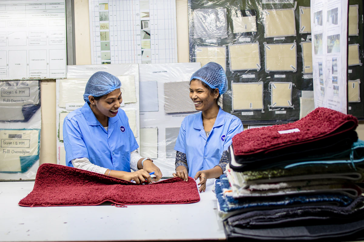 Two women in a textile factory laughing as they check a red rug against specification sheets pinned to the wall behind them