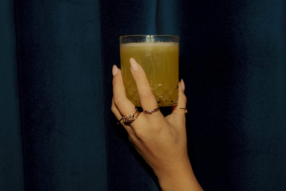A hand holding a glass of freshly pressed juice against a dark curtain