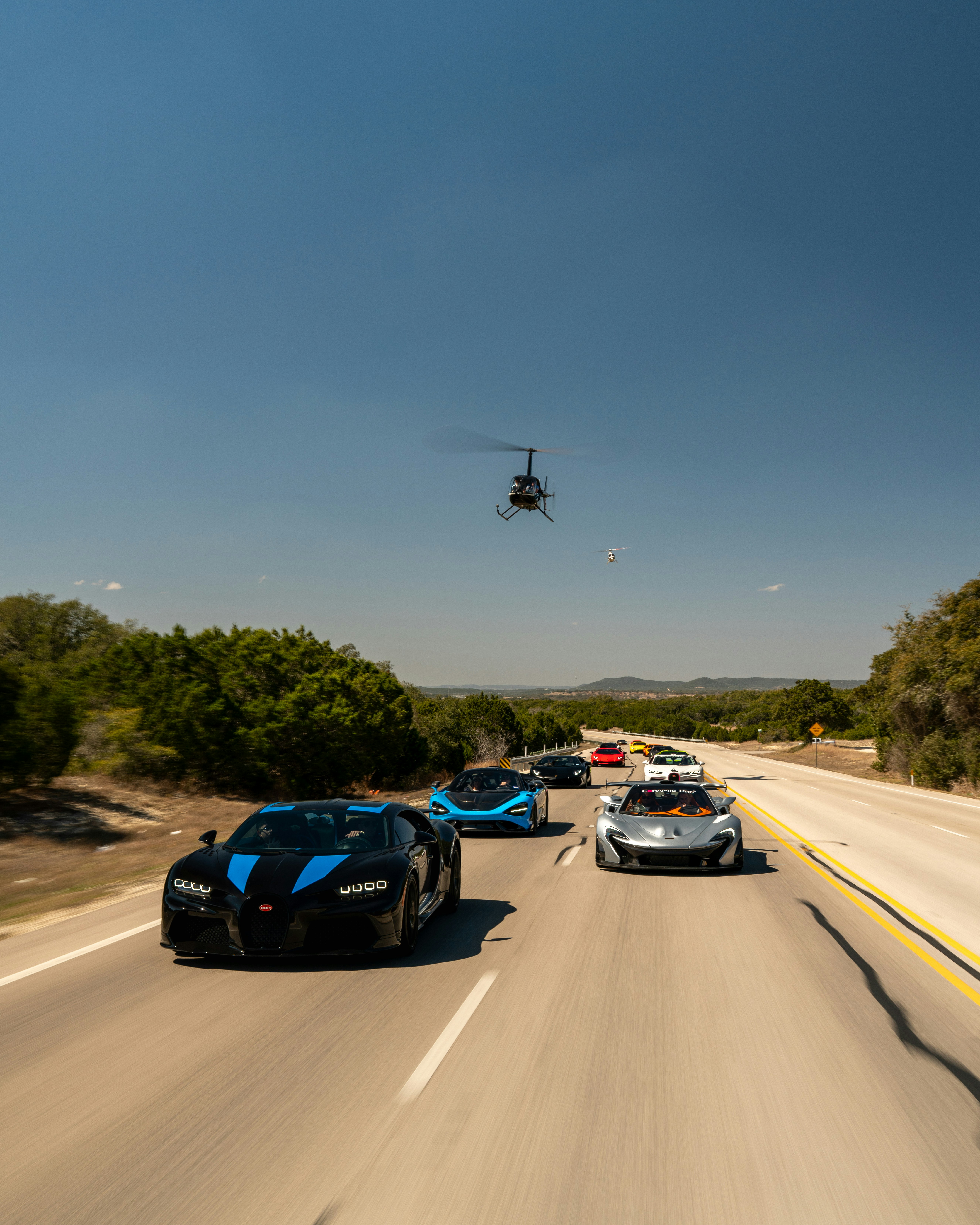 Luxury cars drive along a road with a helicopter.