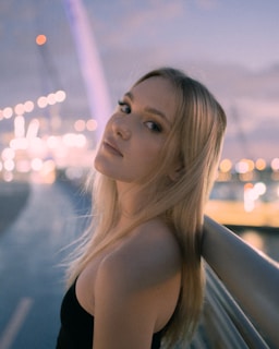 Blonde woman poses by a bridge at dusk.