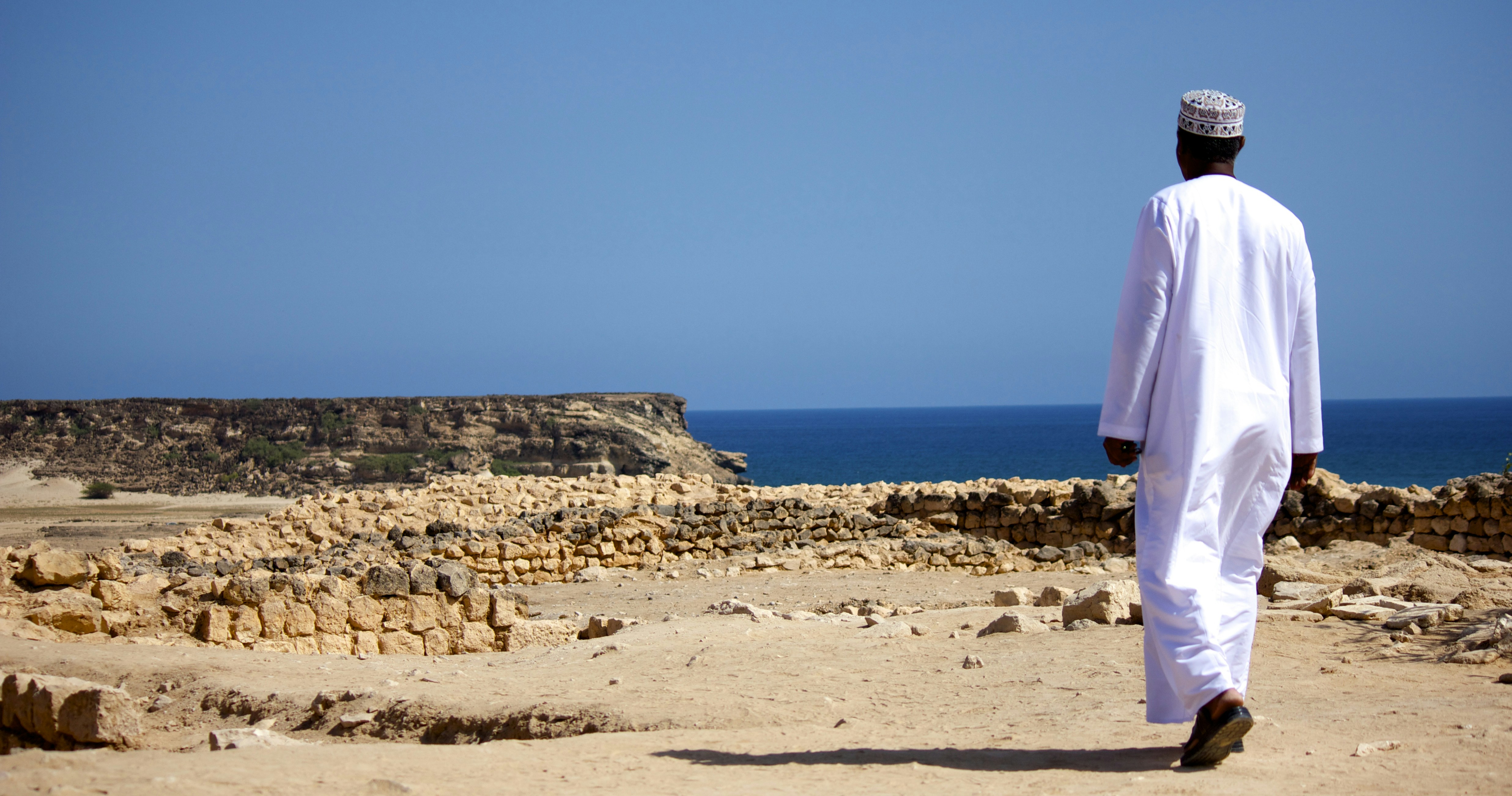 Man walks towards the ocean in traditional attire., Walking with the Wind – Salalah, Oman A man dressed in traditional Omani attire walks through ancient ruins toward the endless blue sea. The wind, the past, and the present all meet here.
