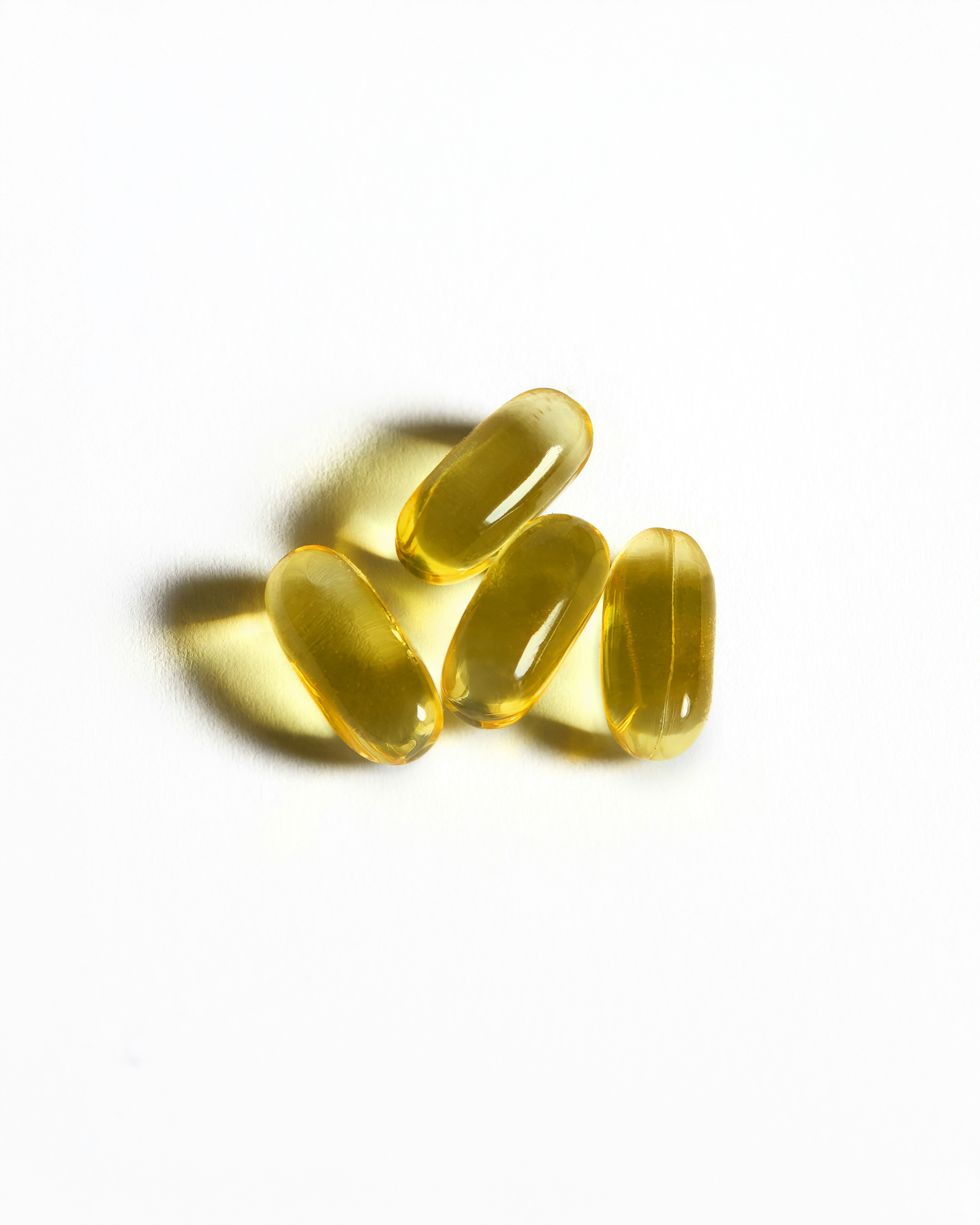 The photograph showcases four golden, translucent Vitamin E capsules placed on a clean white background. The soft lighting highlights their glossy texture, creating an impression of purity and wellness. Vitamin E is known for its powerful antioxidant properties, promoting skin hydration, reducing signs of aging, and protecting against environmental damage. The image conveys a message of self-care and natural beauty.