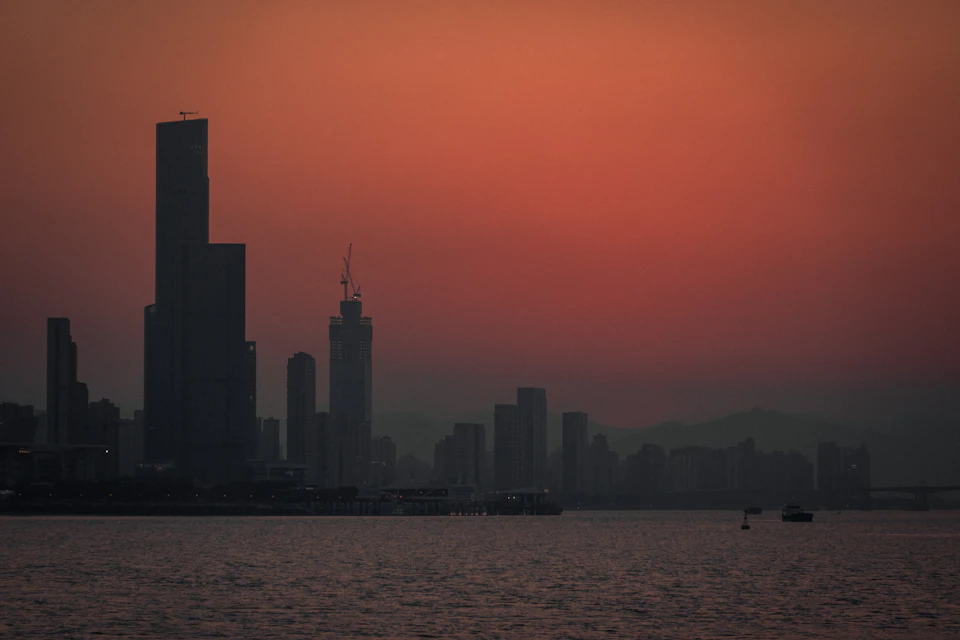 The image depicts a city skyline at sunset, with the sun setting behind the buildings and casting an orange glow across the sky. The tallest building is located on the left side of the frame, while the shortest one is on the right. In the foreground, there are two boats floating in the water near the shoreline, adding to the serene atmosphere of the scene.