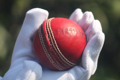 A red cricket ball held in a white glove.