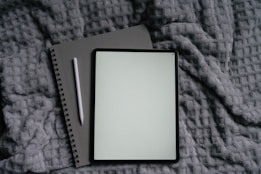 Tablet, notebook, and pen on a gray blanket.