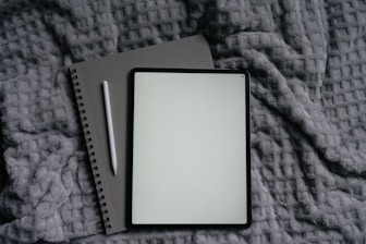 Tablet, notebook, and pen on a gray blanket.