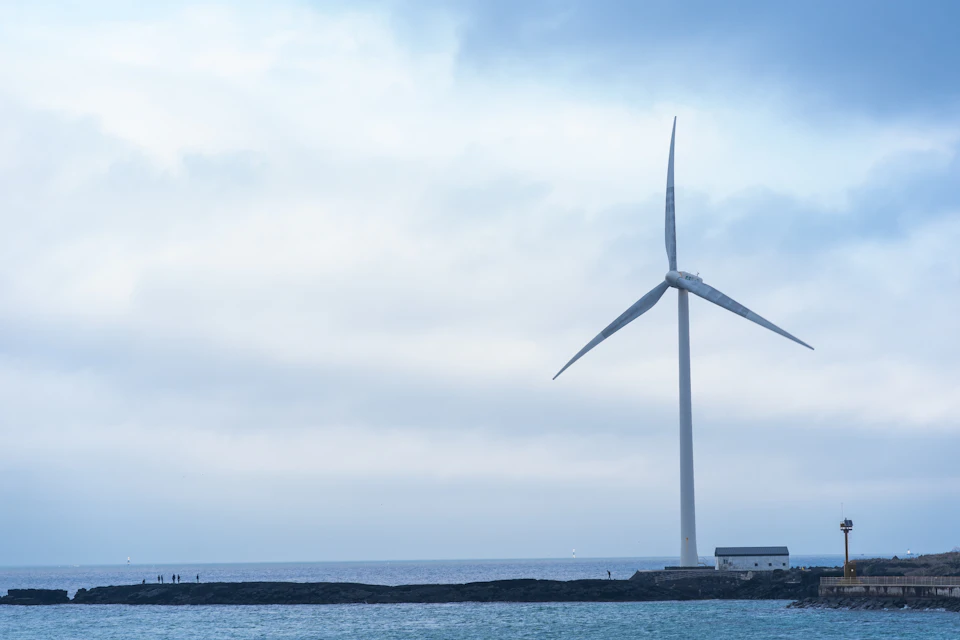 Large white wind turbine stands tall on rocky outcropping overlooking ocean with scattered people
