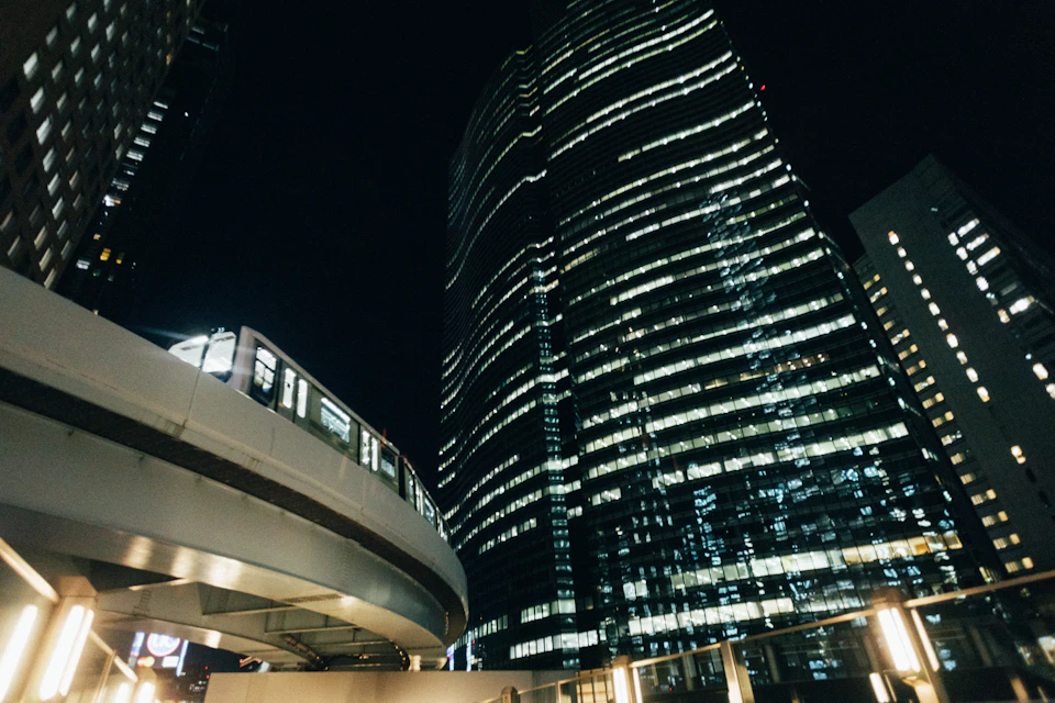 The image depicts a cityscape at night, with a tall skyscraper dominating the scene. The building's facade is illuminated by numerous lights, creating a striking contrast against the dark sky. Below the skyscraper, a train can be seen moving along its tracks, adding an element of modernity to the urban landscape. The perspective of the image is from below, looking up at both the towering structure and the passing train, emphasizing their height and prominence in the city's skyline.

The scene is bathed in the soft glow of streetlights, casting long shadows and highlighting the architectural details of the skyscraper and the train tracks.