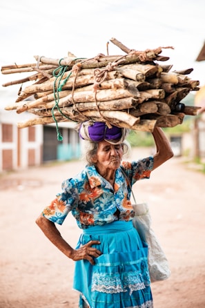 An elderly woman carries firewood on her head.