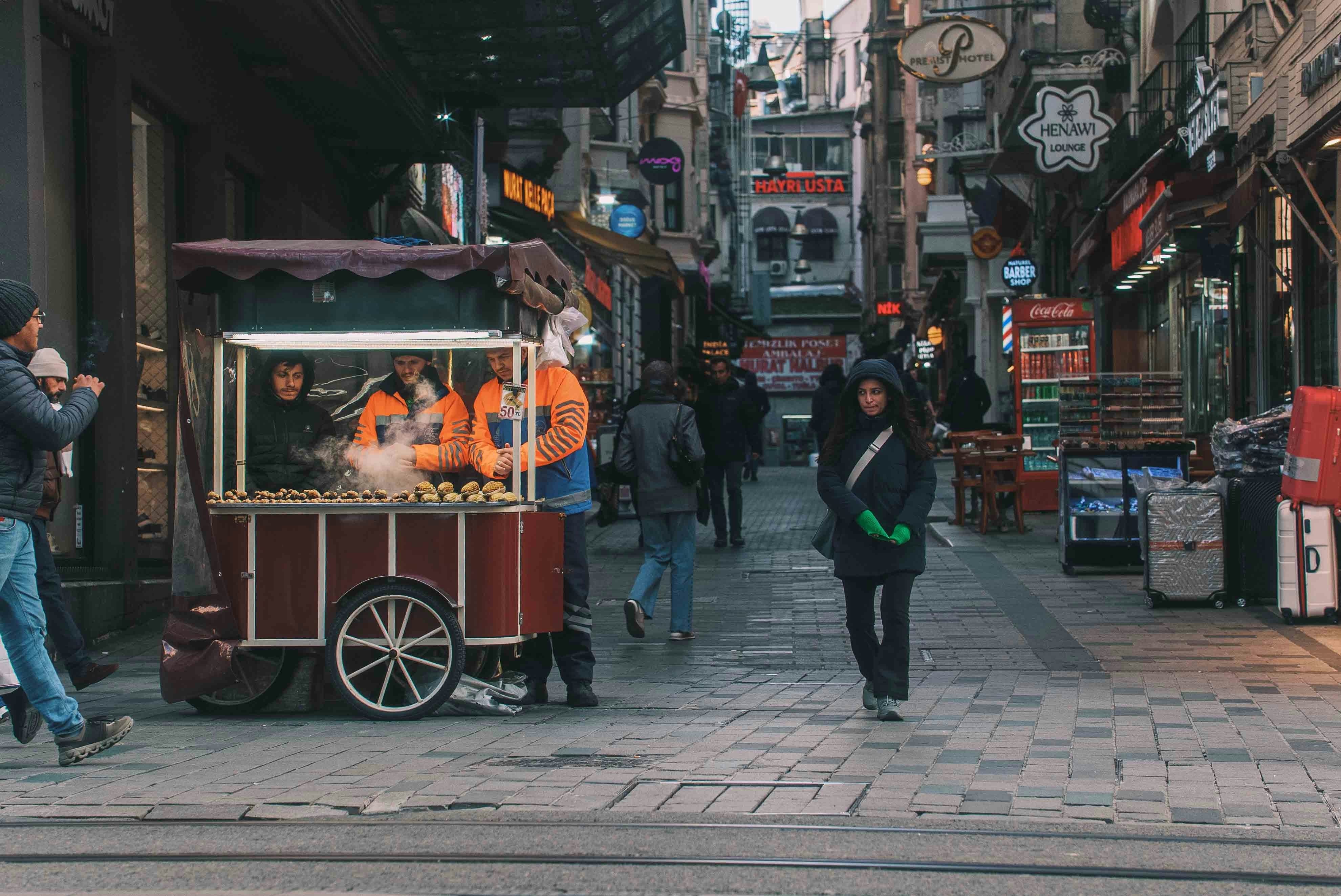 People walk down a bustling street with a food cart.