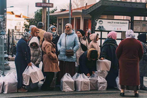 Women wait at a stop with groceries.