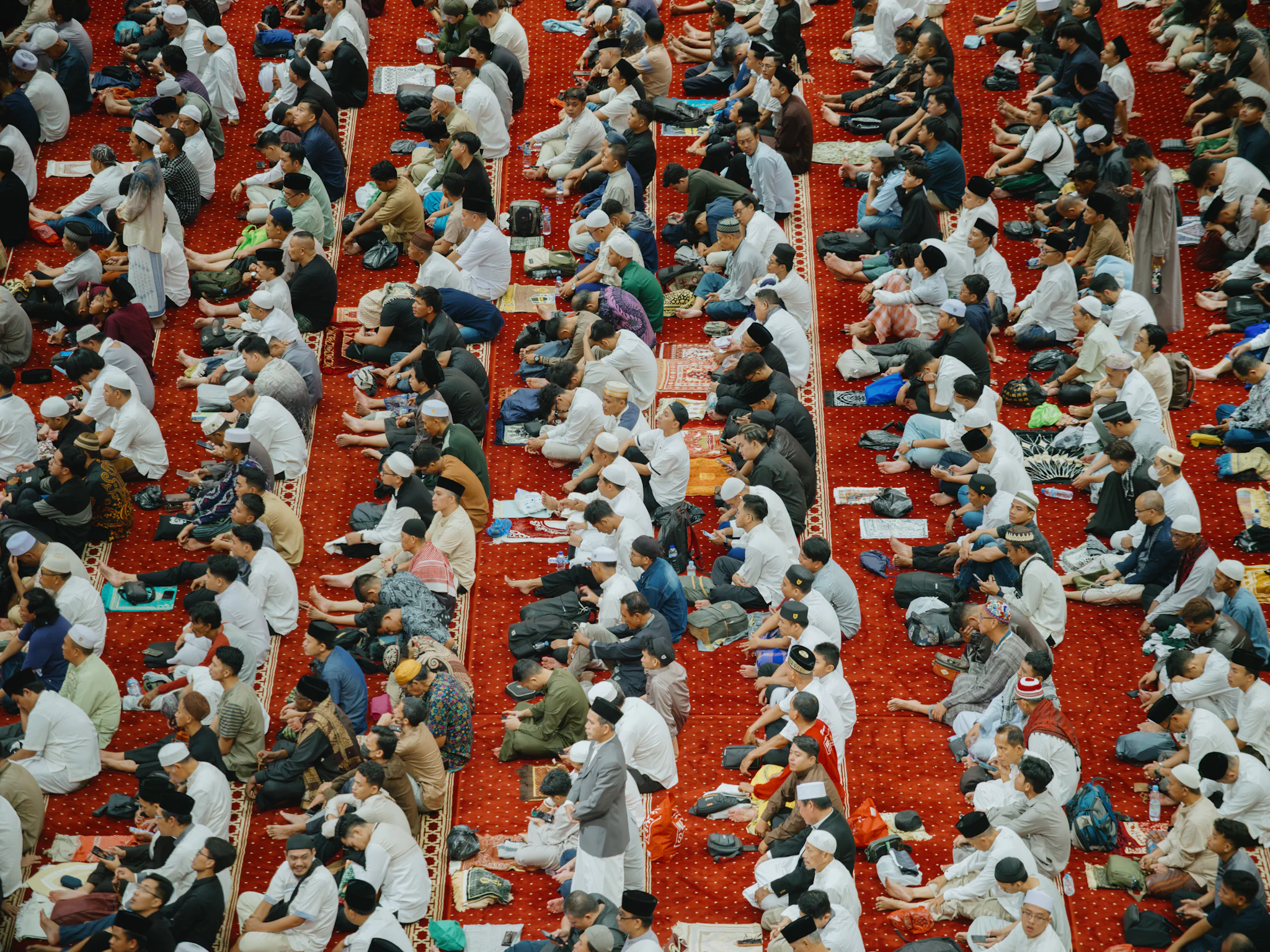 Rows of worshippers seated on red prayer carpets inside a large mosque, seen from above, before Eid al-Fitr prayers