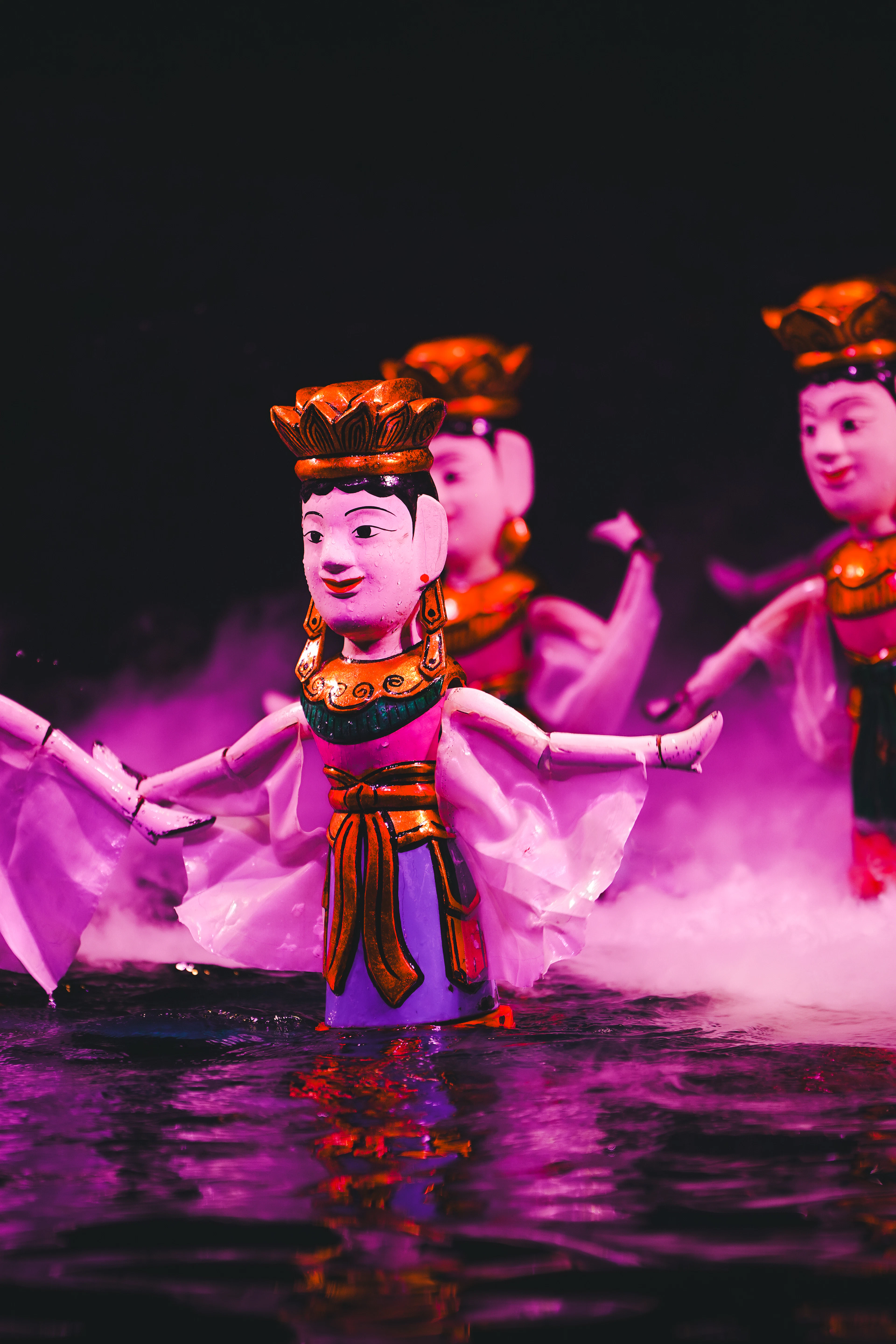 Brightly painted Vietnamese water puppets in lotus crowns and pink robes dancing on the surface of the water in magenta stage light and drifting mist at a Hanoi water puppet show.