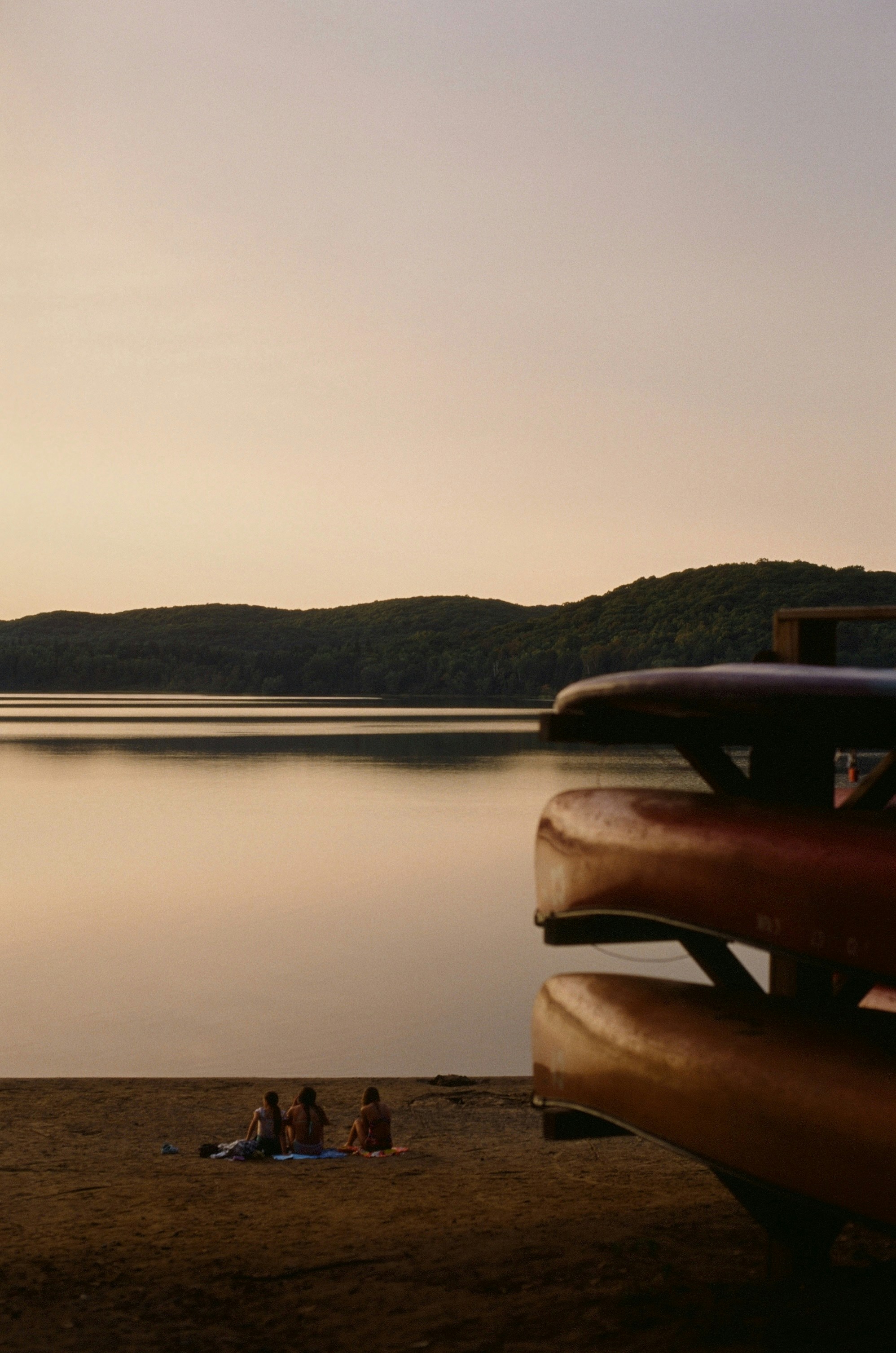 People relax by a lake near canoes at sunset.