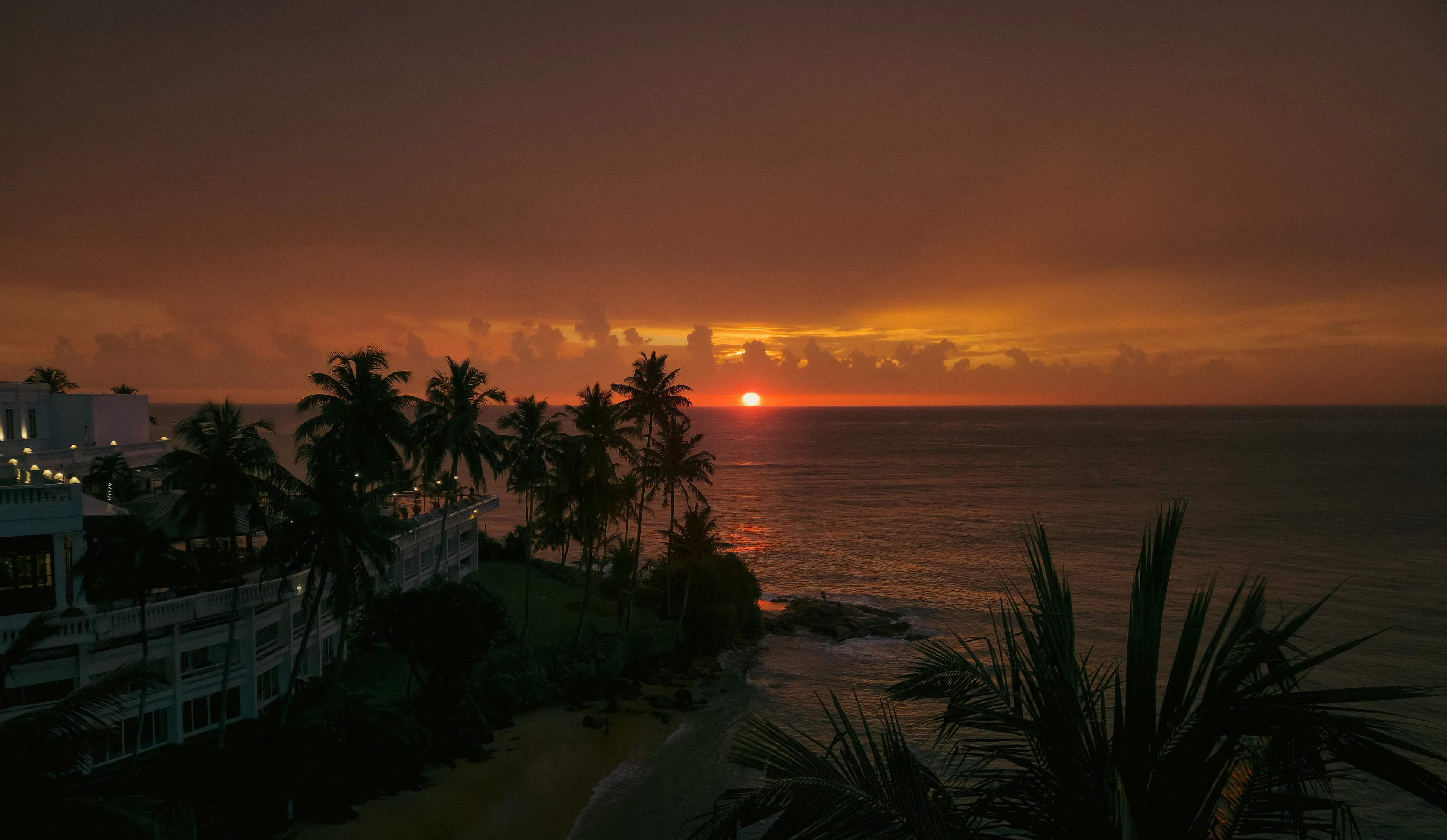 A fiery golden sunset over the ocean with a beachfront hotel and palm trees in silhouette.