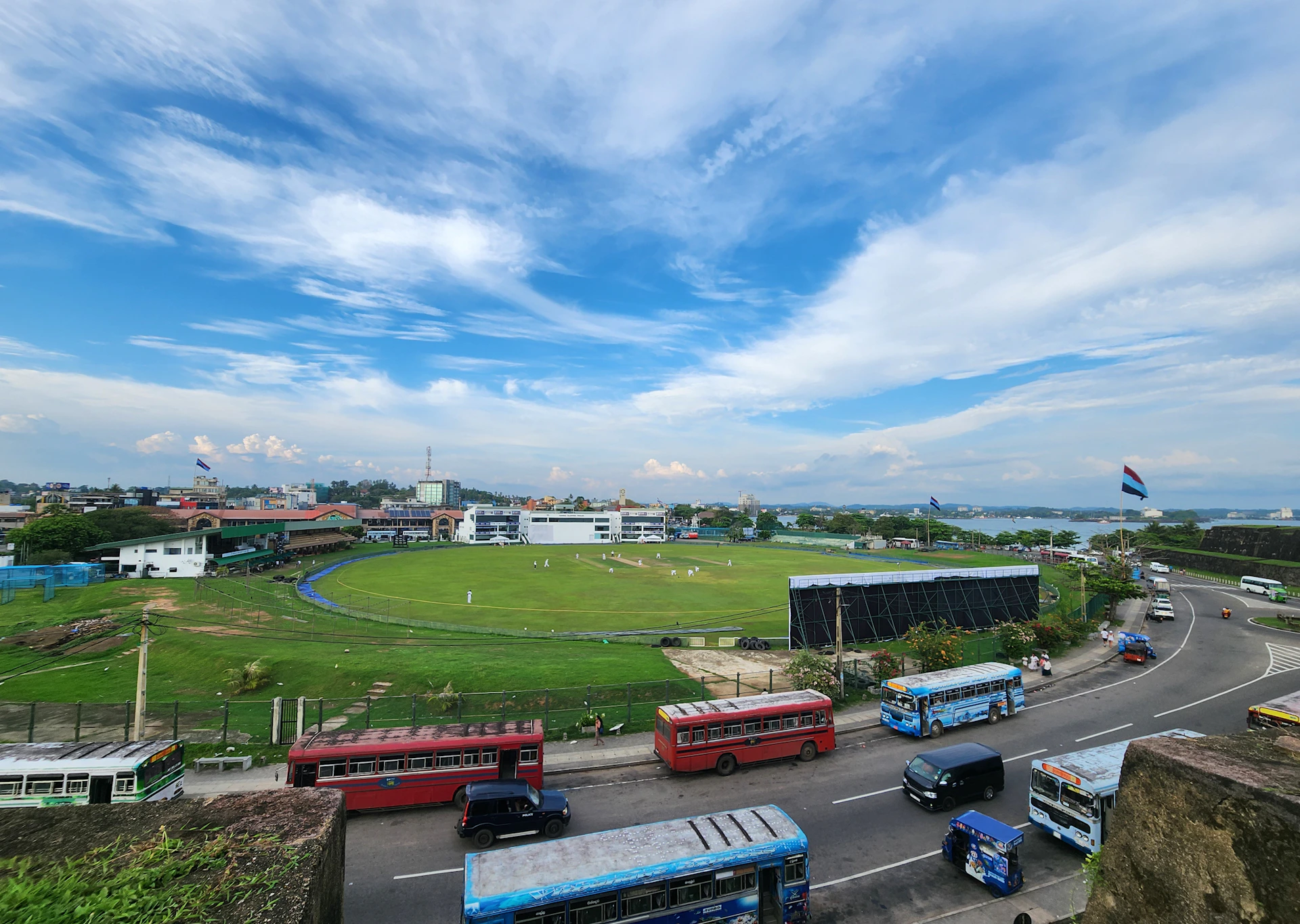 Buses drive along a road near a large, grassy field.