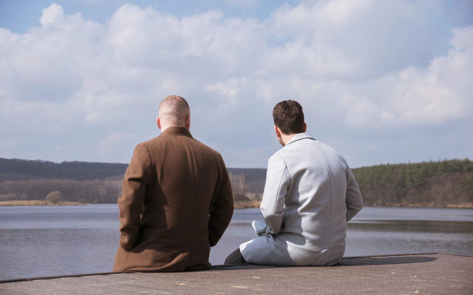 A father and his grown son sitting side by side on a lakeside pier, looking out at the water