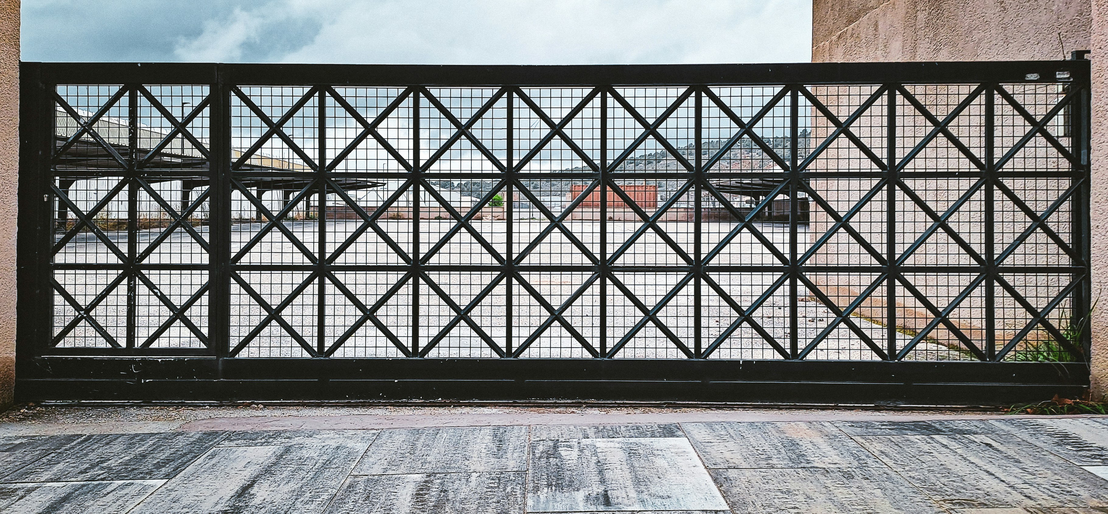 Closed black rectangular metal gate to limit access to car parking area in open space with shelter on left and mountains under cloudy sky in distance.
