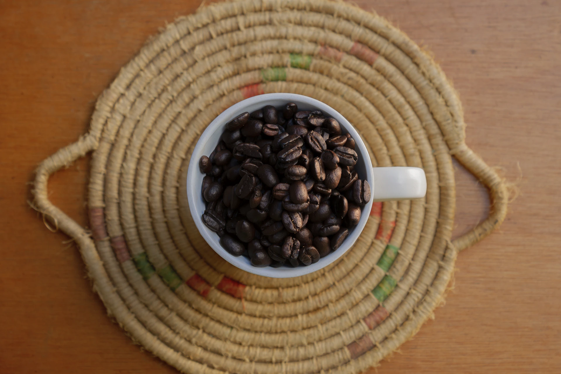 Coffee beans fill a white cup on a woven mat.