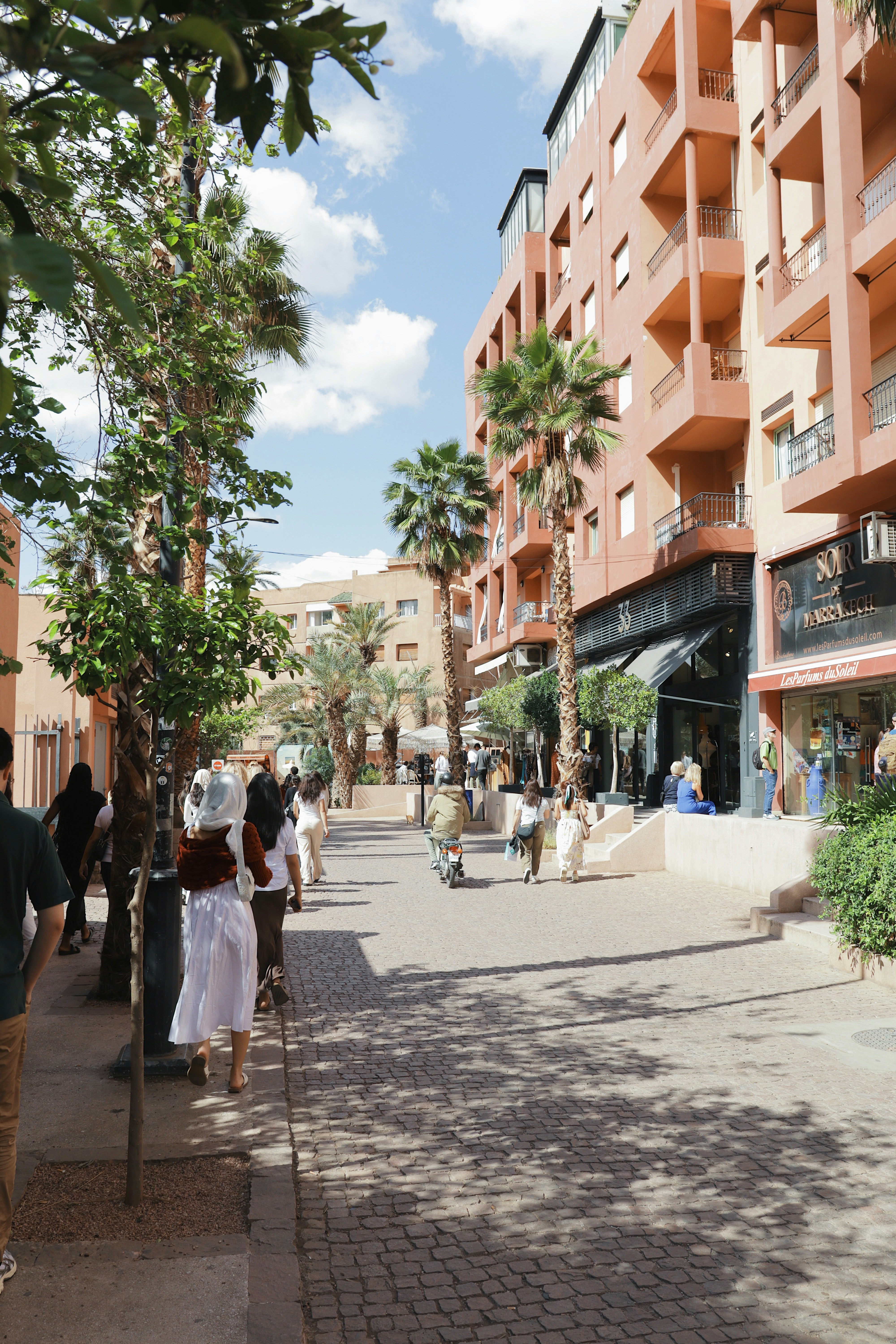 People stroll down a sunny street with buildings.