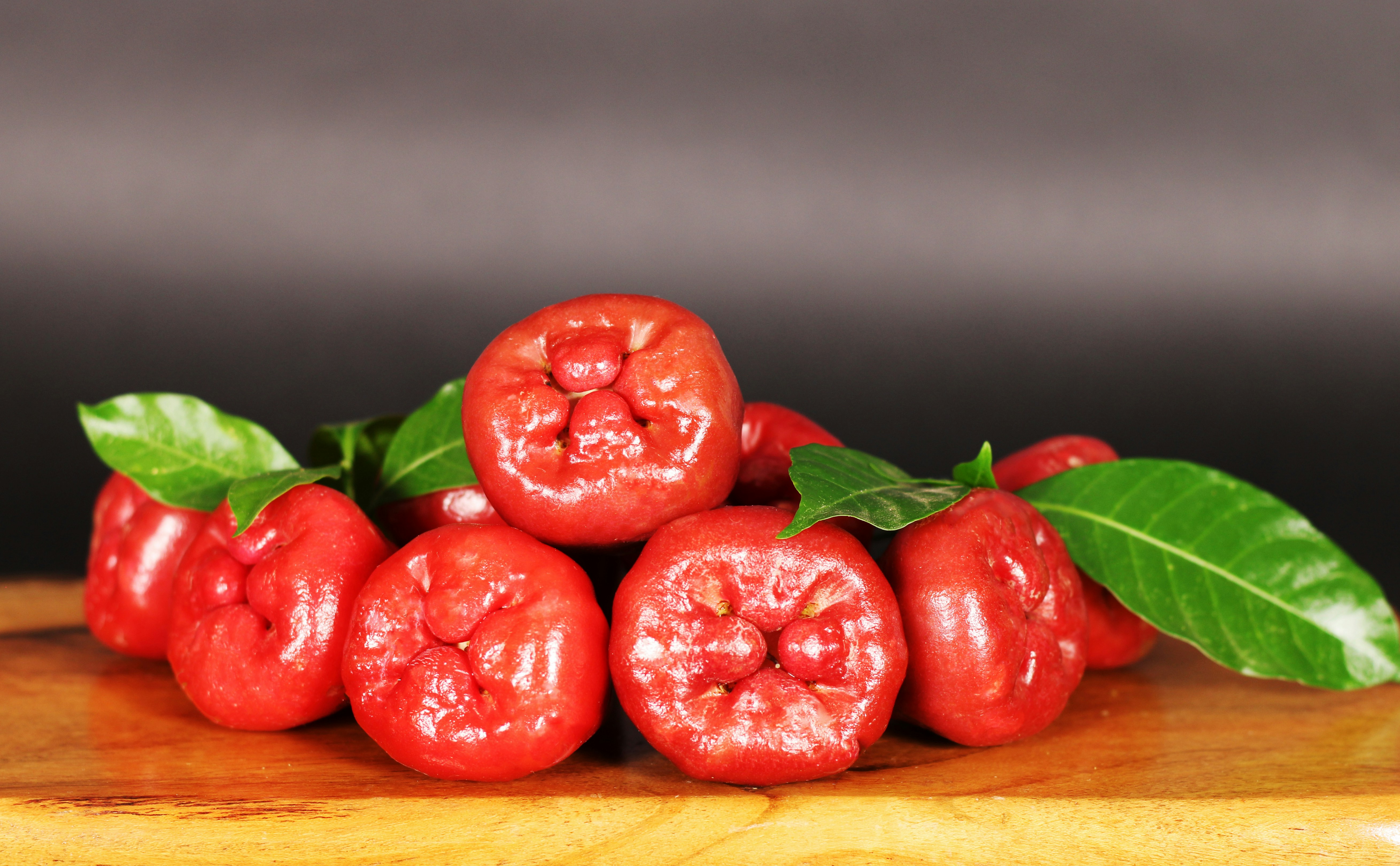 A vibrant cluster of ripe rose apples, freshly picked and displayed on a rustic wooden surface. Their glossy red skin and lush green leaves capture the essence of tropical freshness and natural beauty.
