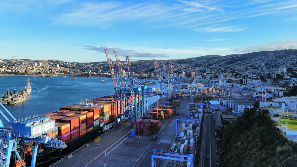 The image presents an aerial view of a bustling port city, with several large cargo ships docked at the harbor and cranes towering over them. The largest ship is positioned on the left side of the frame, while smaller vessels are scattered across the right side. The harbor itself is a hive of activity, with numerous cranes and other equipment visible in various stages of operation. In the background, the cityscape unfolds, featuring buildings of different sizes and designs that add depth to the scene. The sky above is a clear blue, dotted with fluffy white clouds, providing a beautiful backdrop for this industrial landscape.