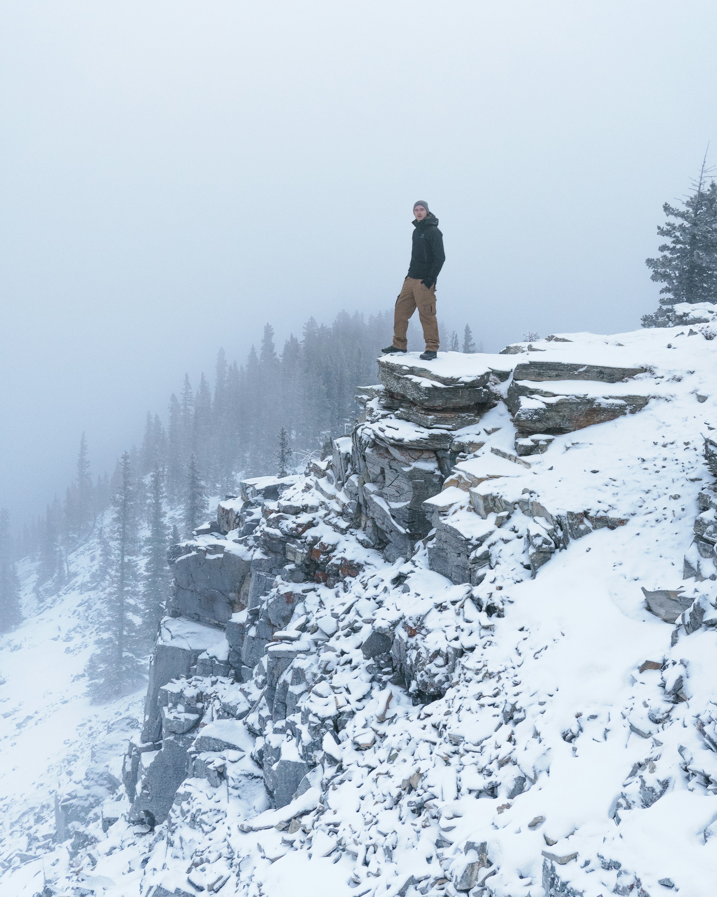Man stands on snowy cliff, overlooking a misty forest.