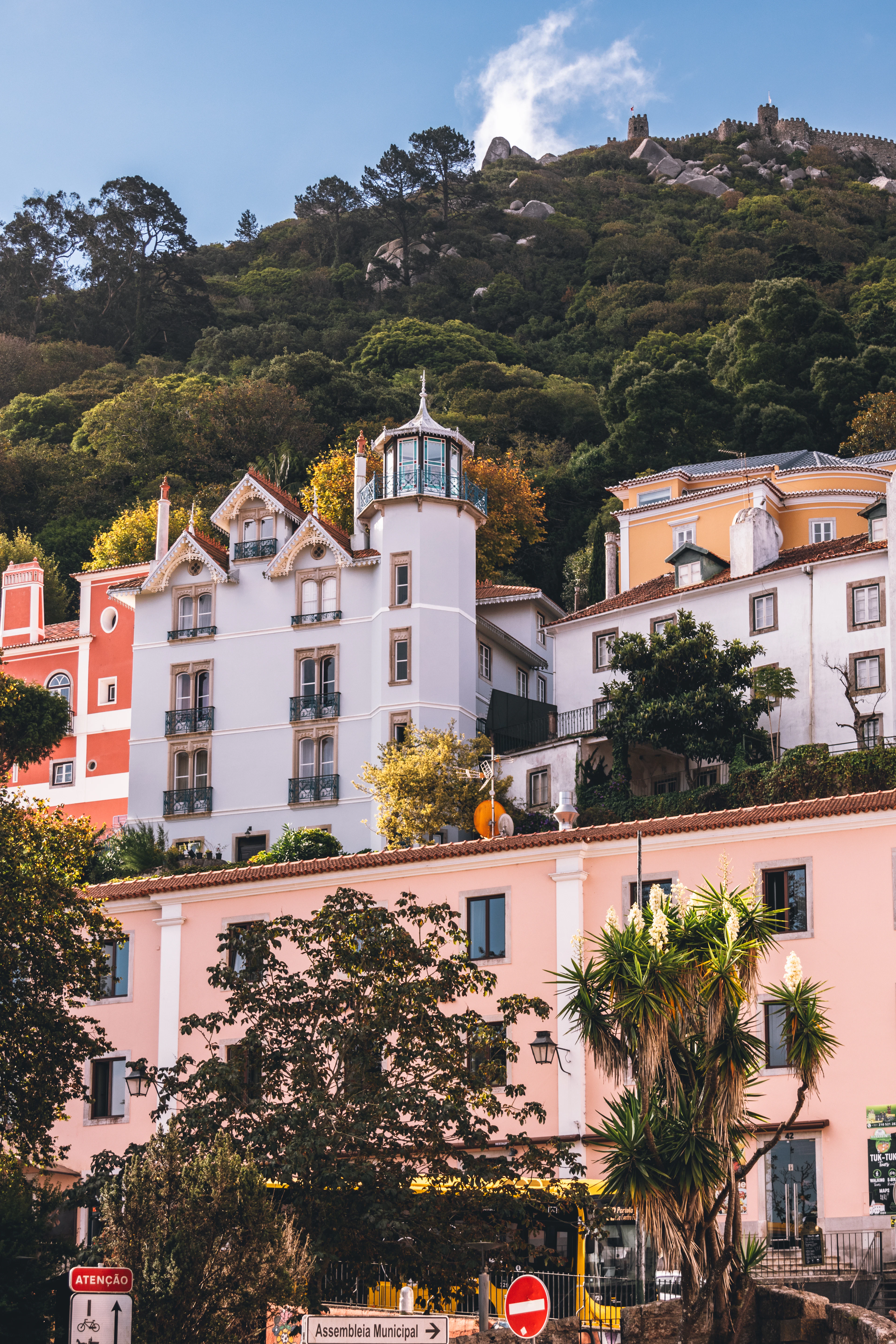 Pastel townhouses of Sintra in coral, cream and pink stacked up a wooded hillside, with the stone ramparts of the Castle of the Moors running along the ridge above them