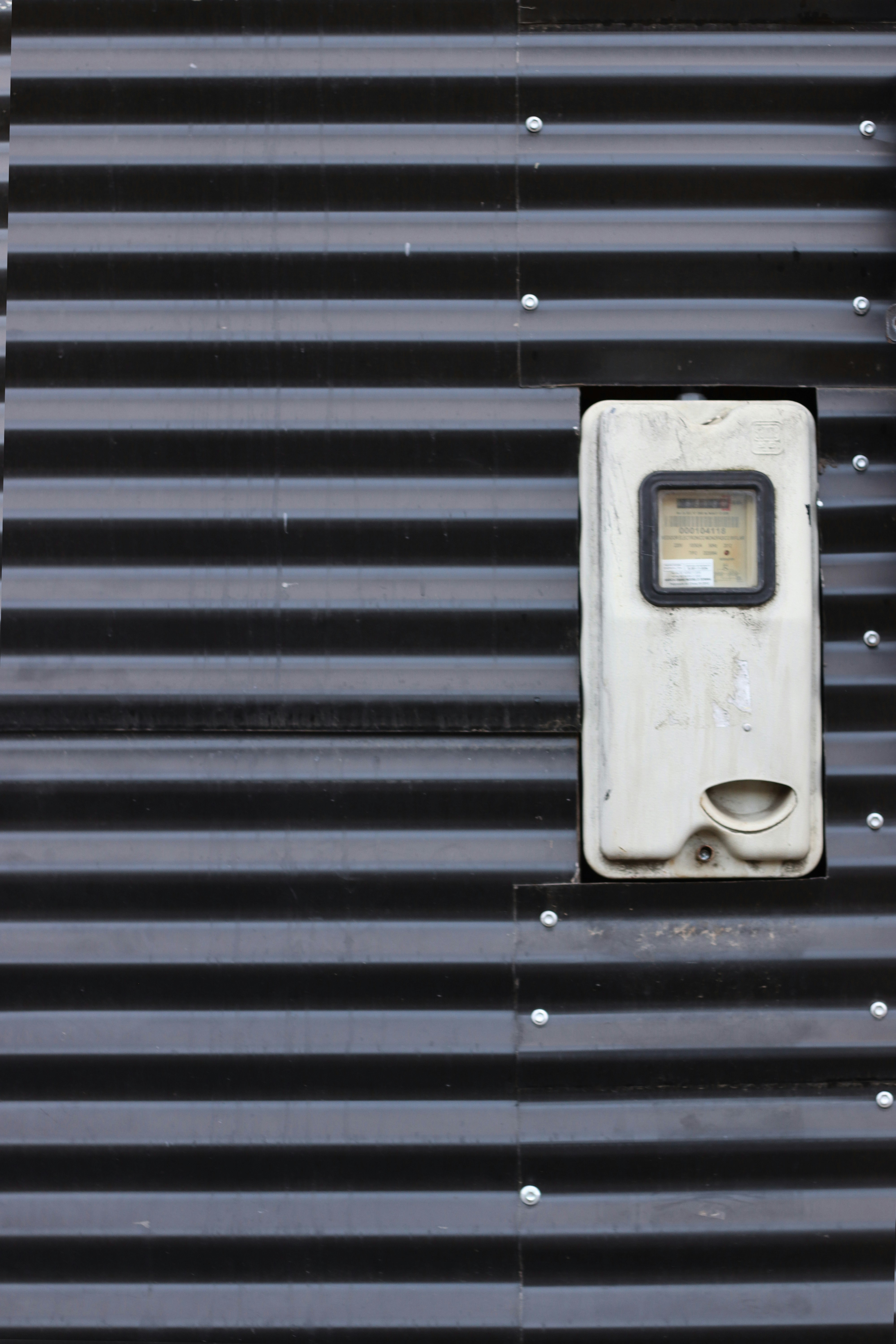 An electric meter sits on a corrugated wall.