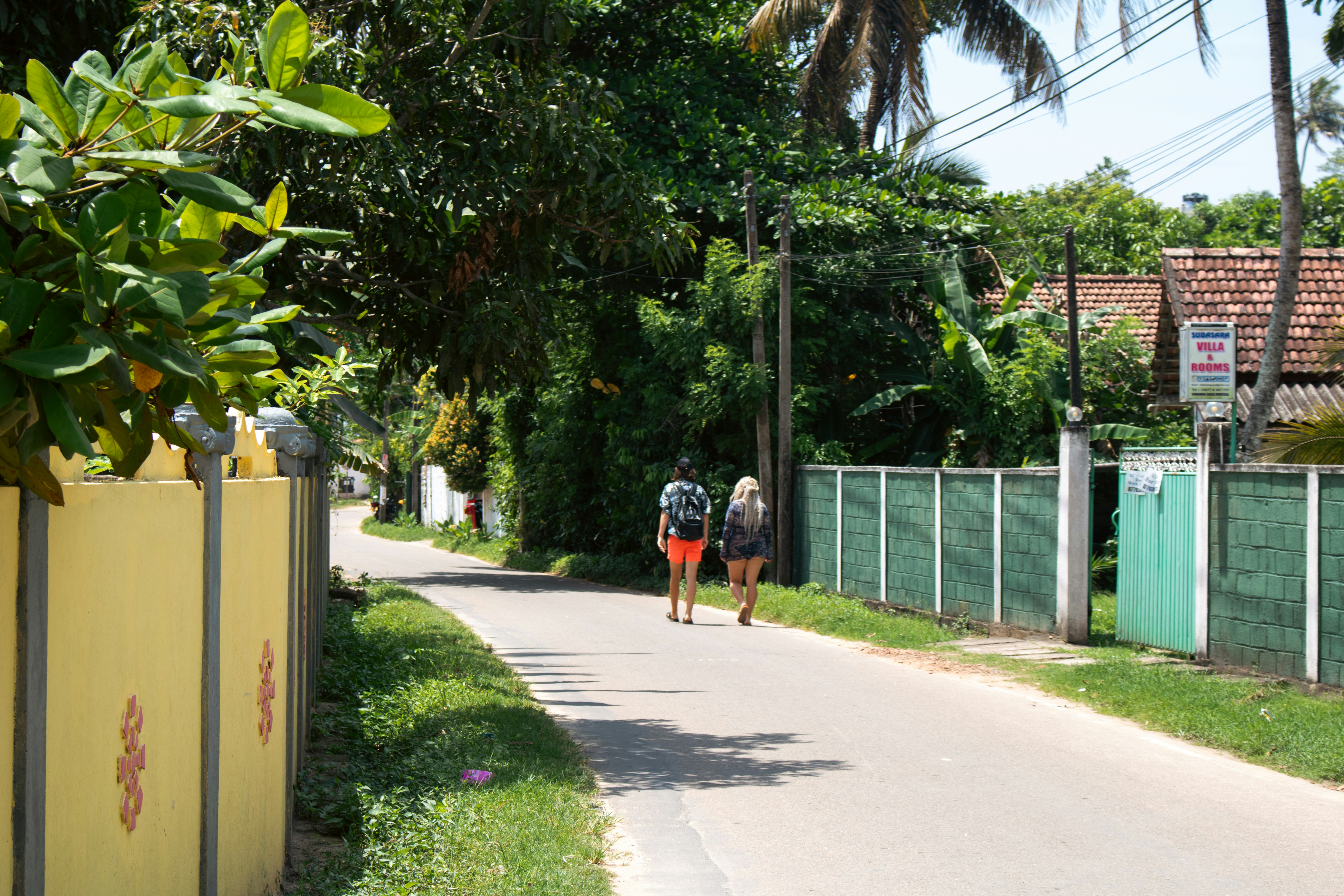 A person in a white tee and shorts walking down a narrow paved street, within Mirissa.