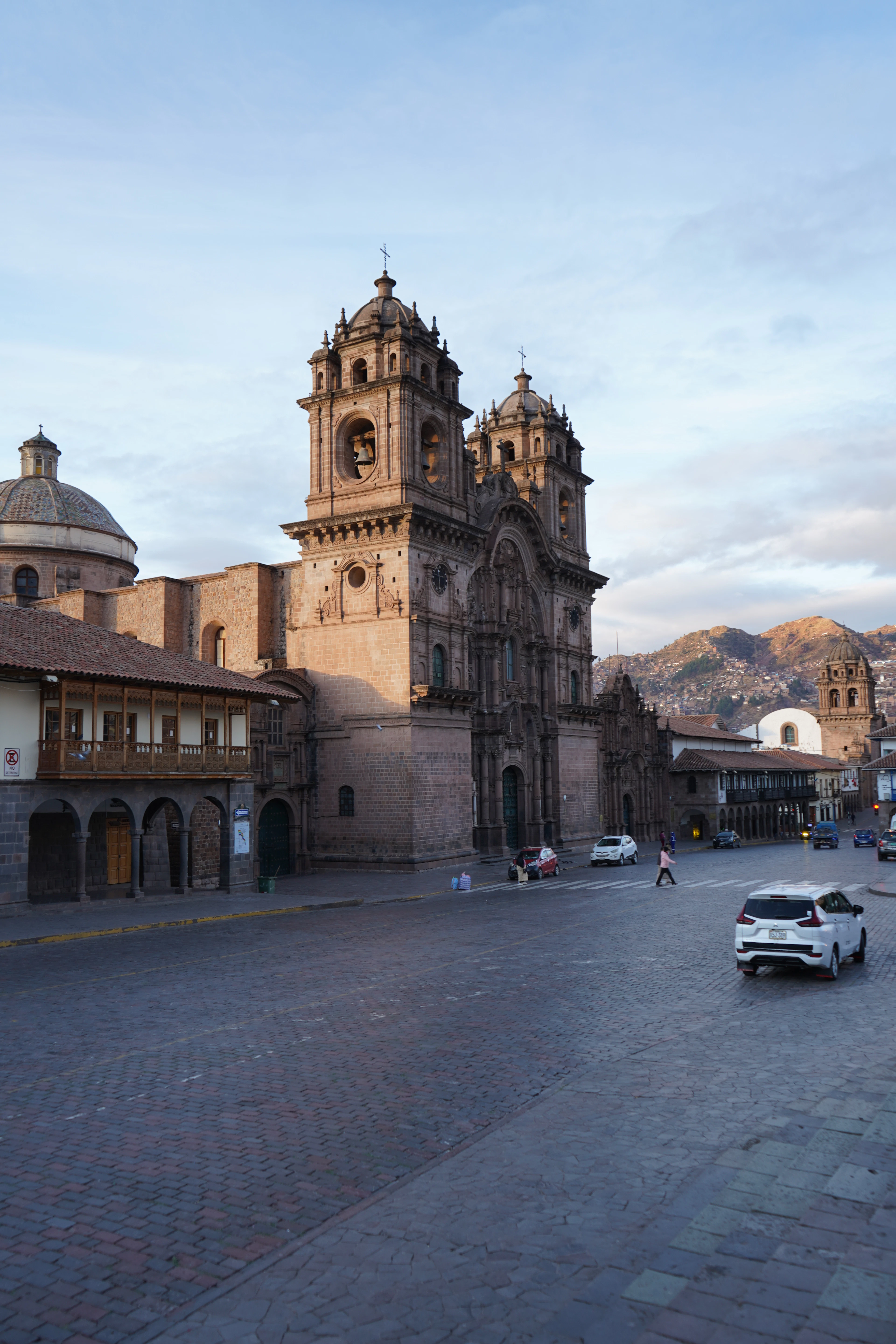 Early light on the baroque towers of the Iglesia de la Compañía de Jesús on Cusco's Plaza de Armas, the cobbled street in front all but empty, green hills stacked with houses rising behind the roofline.